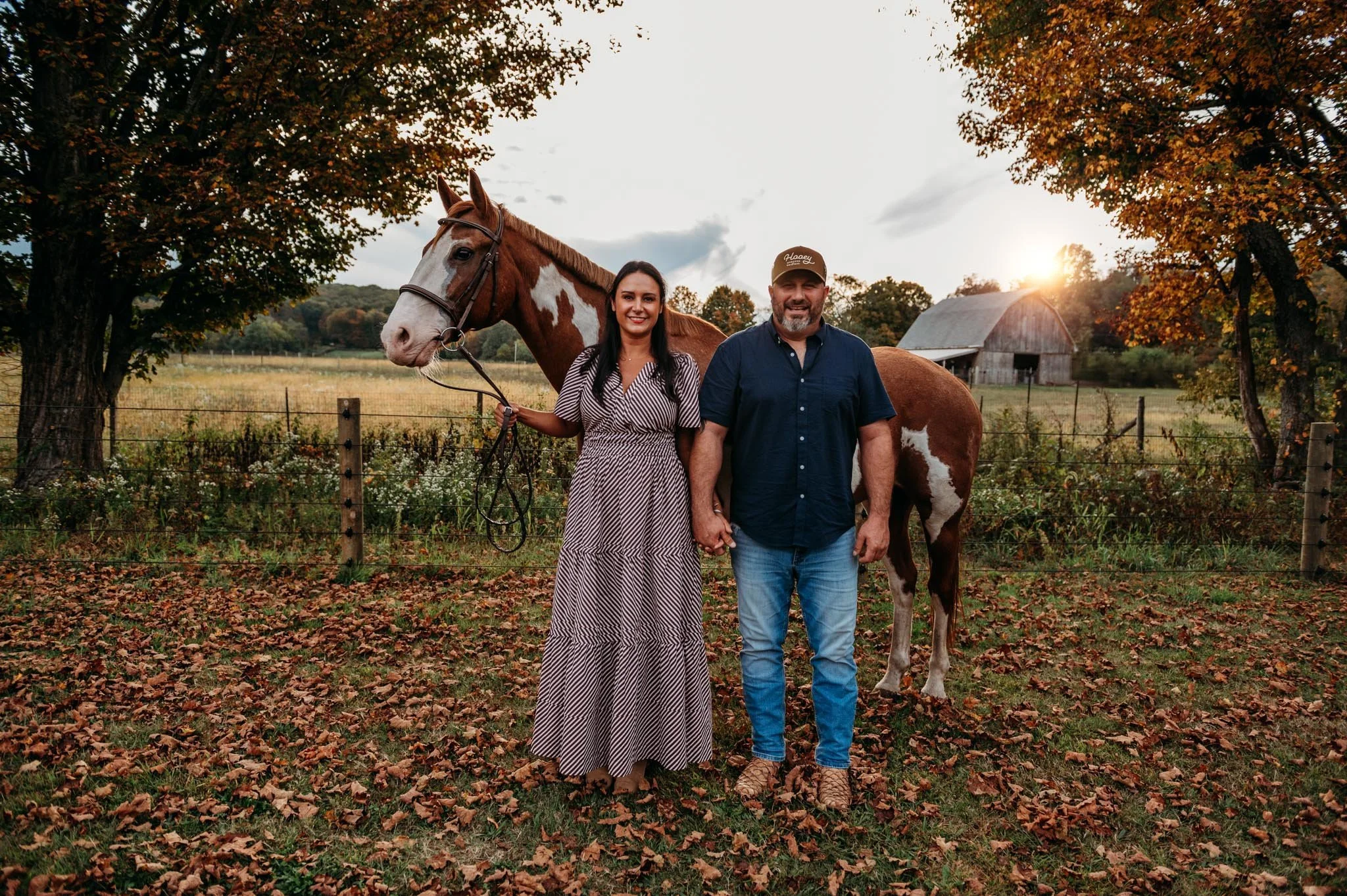 A woman and a man standing hand in hand beside a brown and white horse in a rural setting with autumn leaves on the ground and trees in the background during sunset connecticut dog breeder cavapoo pupppies for sale