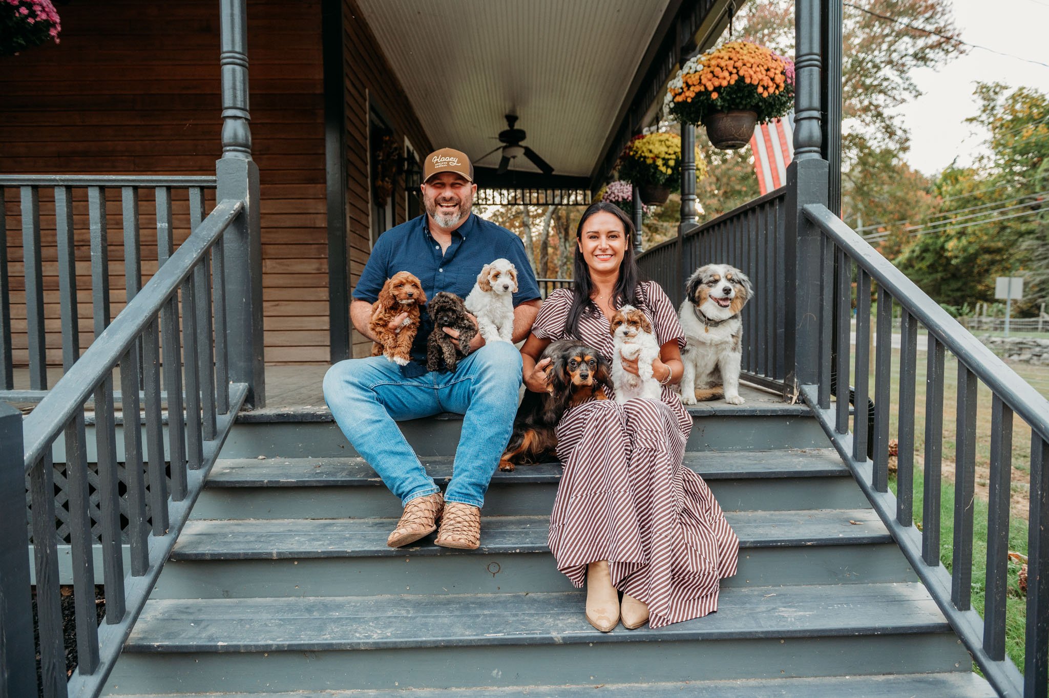 A man and a woman sitting on the steps of a porch holding puppies, with more dogs sitting beside them, house and trees in the background cavapoos for sale dog breeder in connecticut