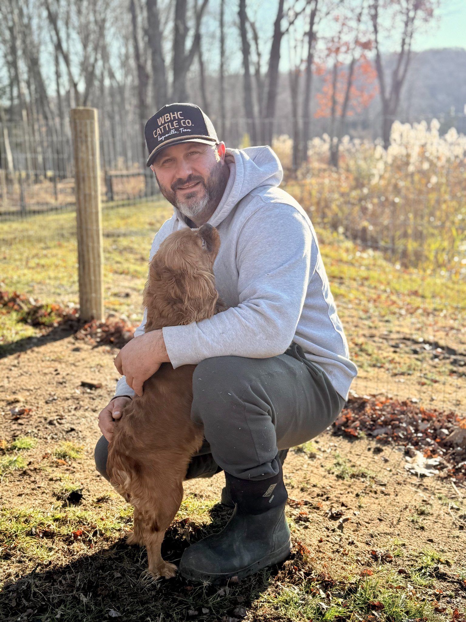 rob in a cleared yard holding a brown puppy with long ears, wearing a gray hoodie, black pants, and black boots, with a background of trees and a sunny sky cavapoo puppies connecticut