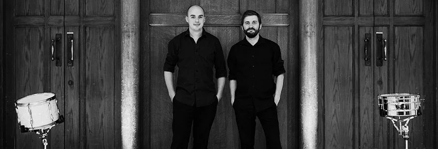 Percussion group O Duo standing in a room with drums against  wood background
