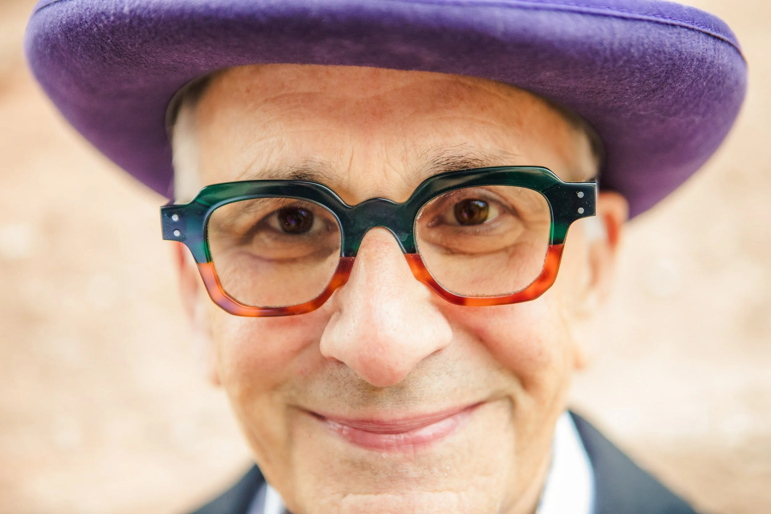 Close-up of a smiling man wearing a purple hat and colorful, thick-framed glasses.