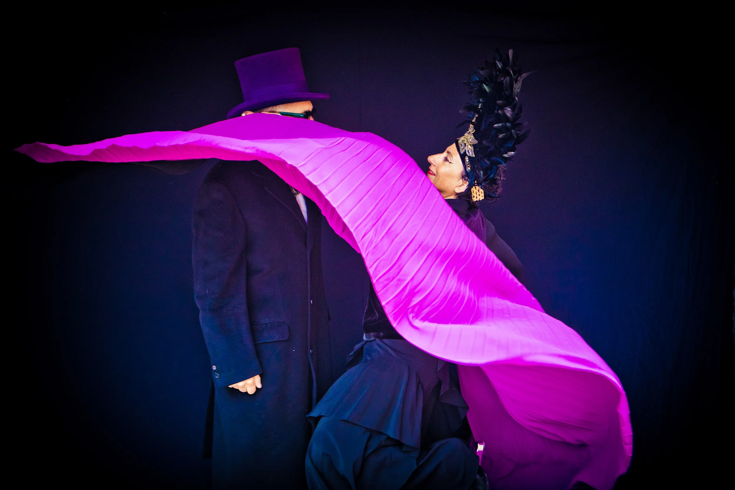 Woman with black feather headdress dancing in front of man in purple top hat, flowing magenta fabric in foreground