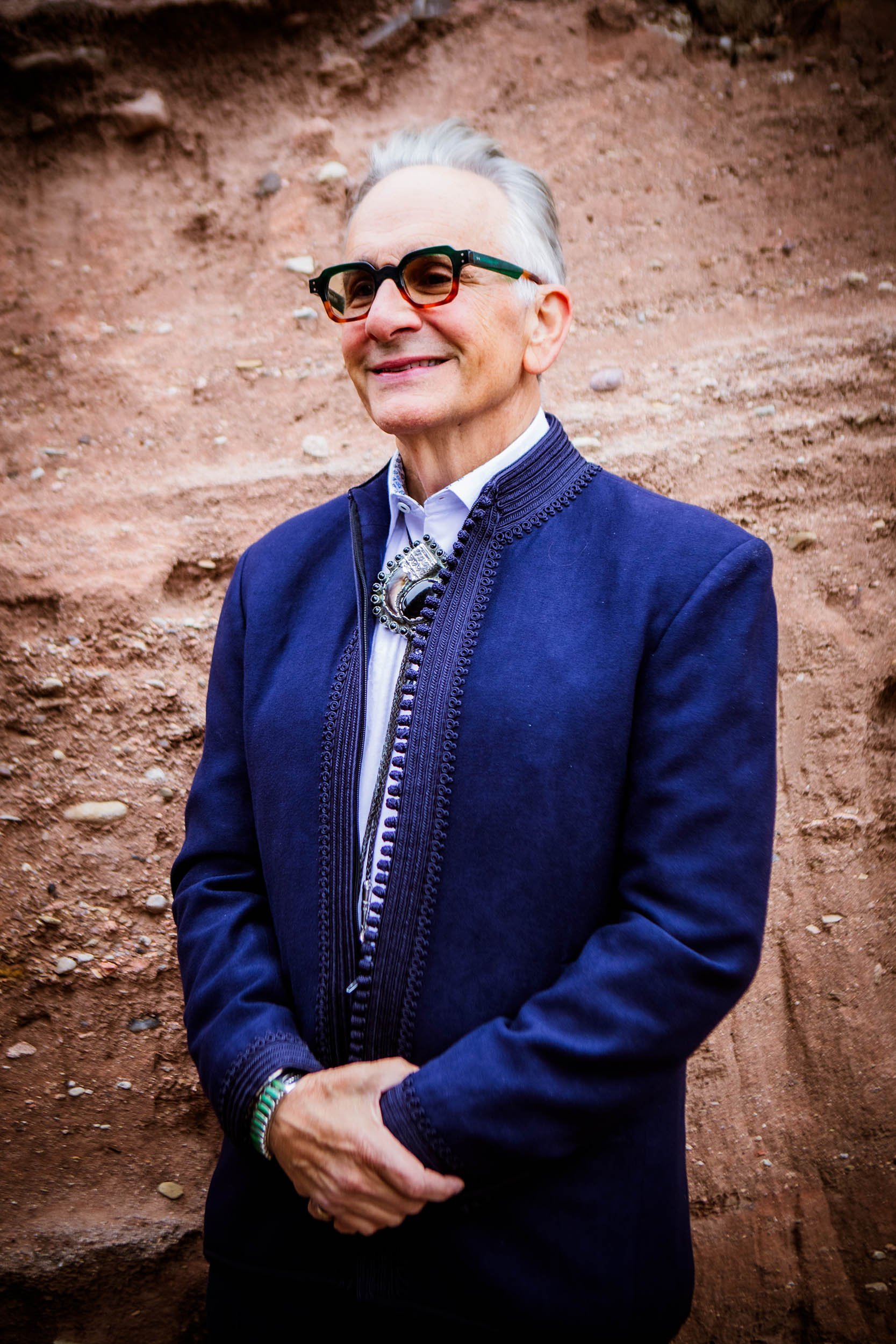 Older man with white hair wearing glasses, blue jacket, button up shirt and bolo tie with hands clasped