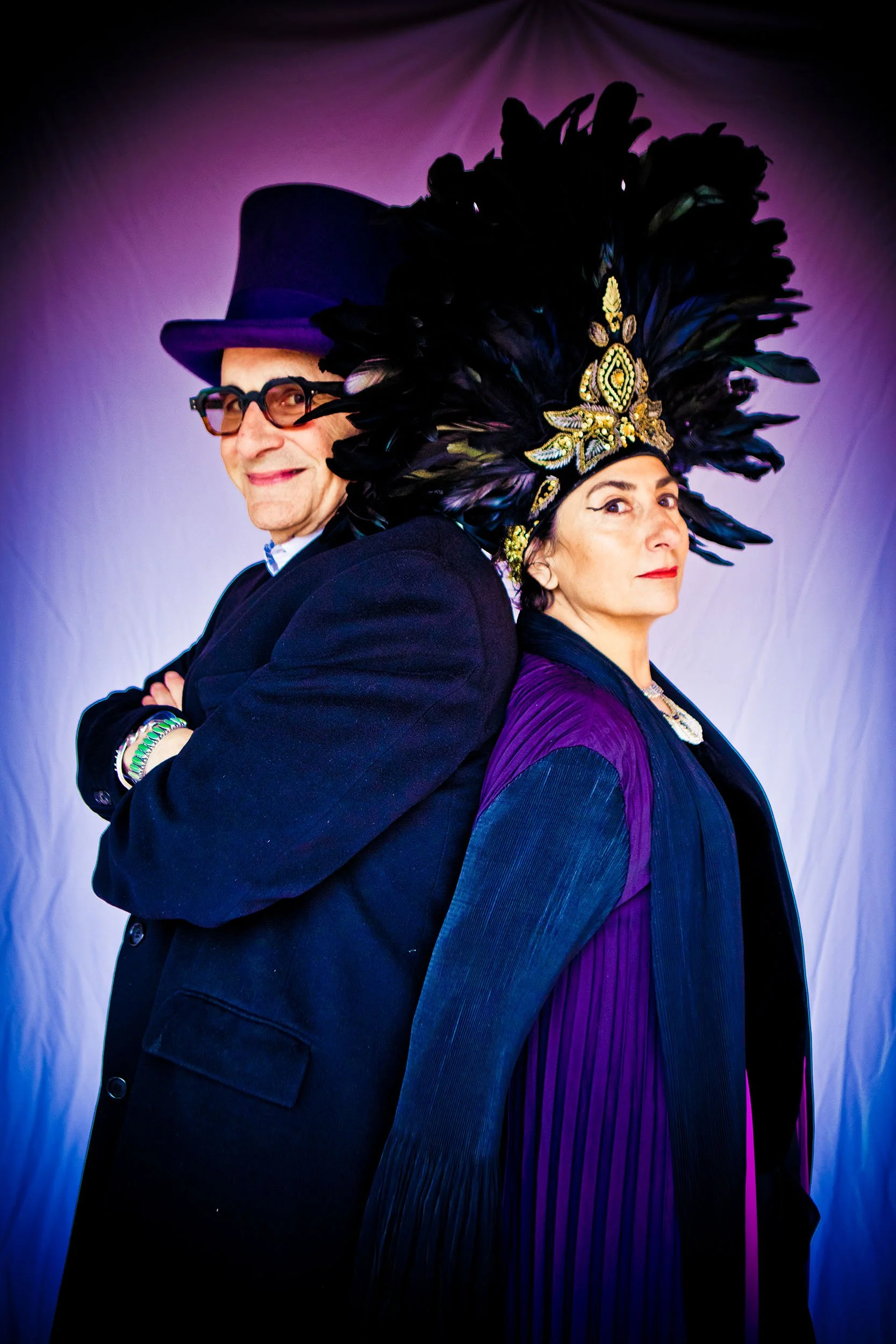 Senior man wearing glasses, purple top hat and long coat with arms crossed back to back with woman wearing black feather headdress wearing blue and purple flowing costume