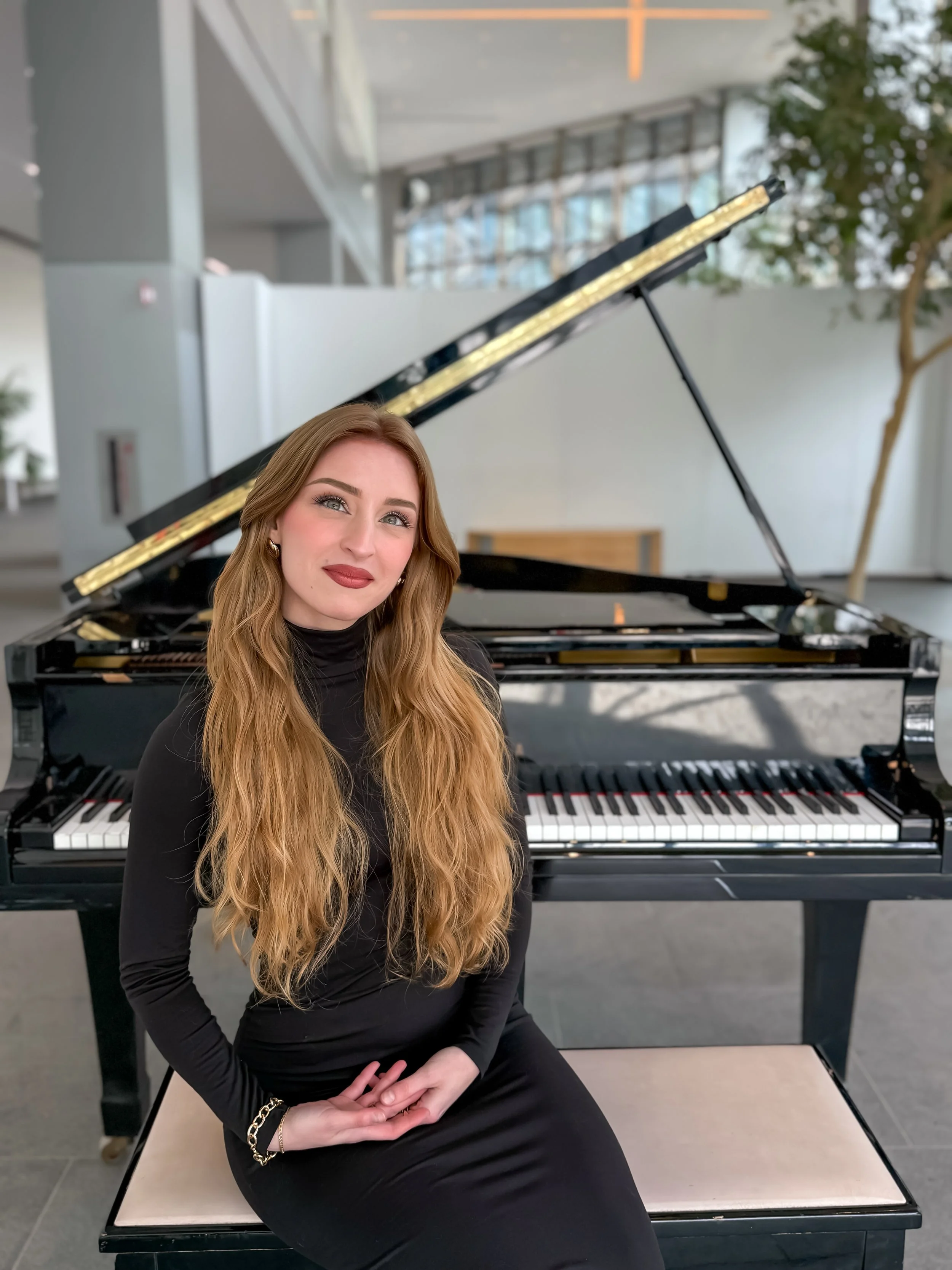 A woman with long, wavy blonde hair in a black dress sitting on a piano bench in front of an open grand piano in a modern, well-lit indoor space.