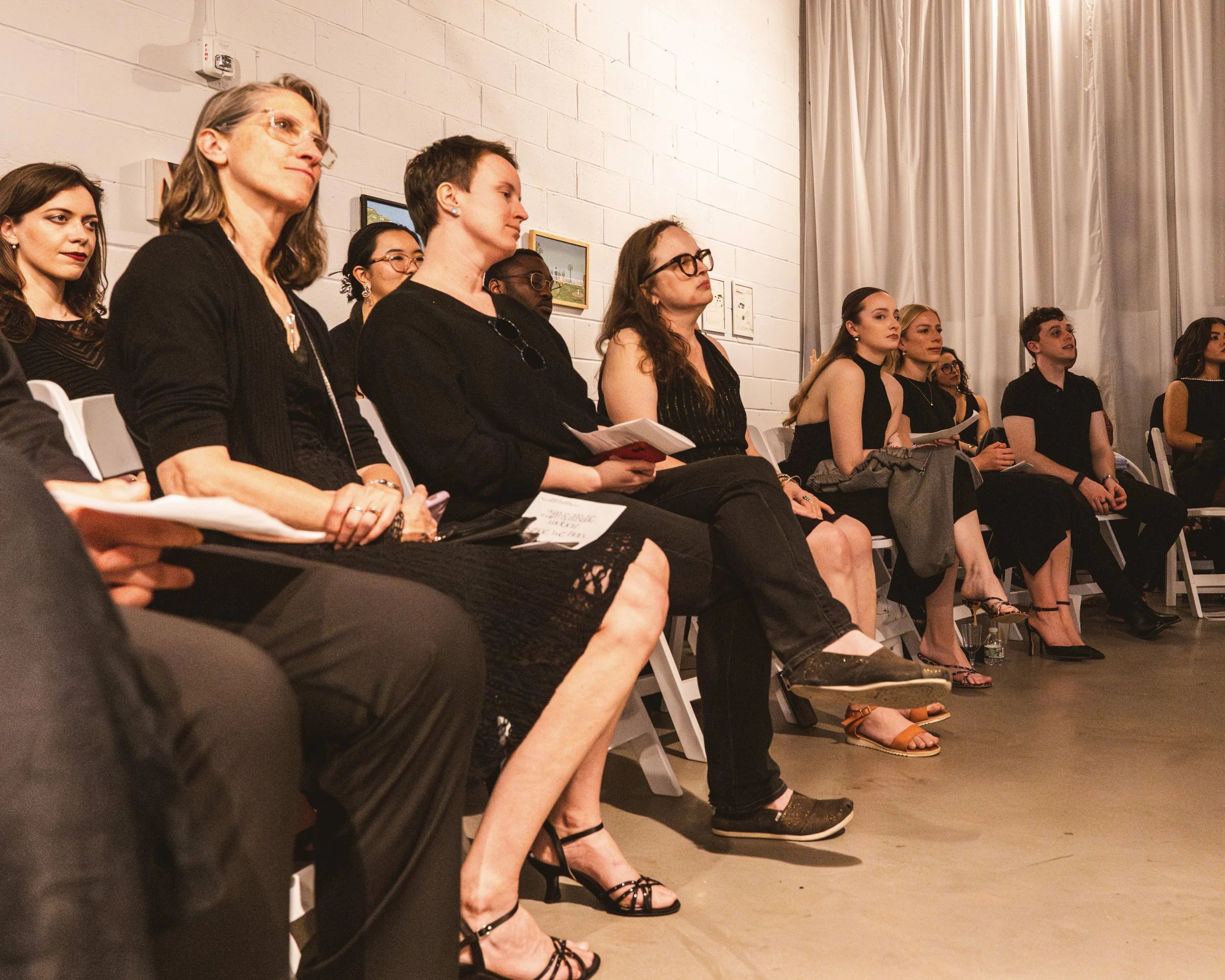A group of people sitting on chairs in a room, attending an event or presentation. The attendees appear focused or listening attentively, with some holding notebooks or papers, and dressed in formal or semi-formal attire.