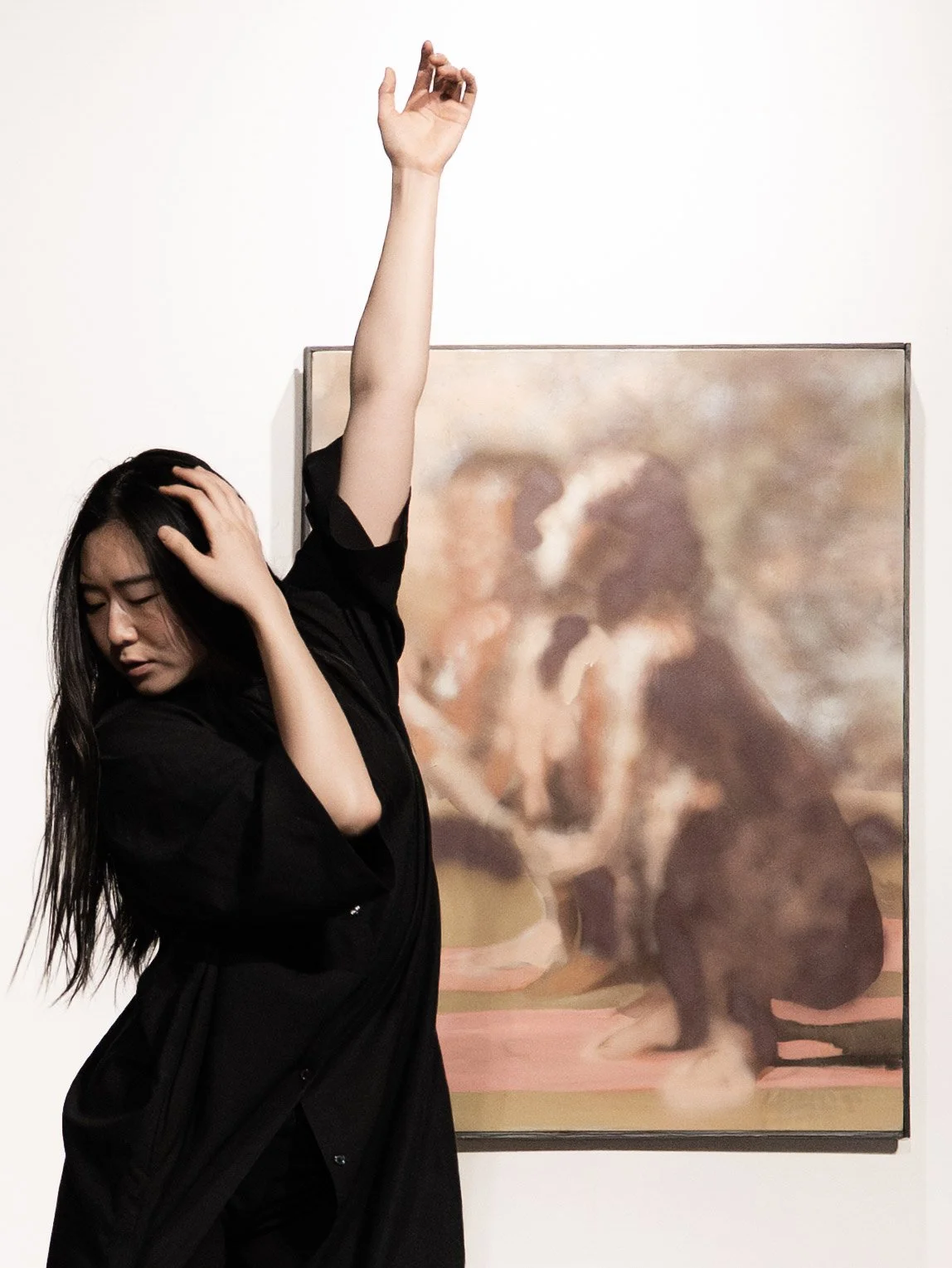 A woman with long black hair in black clothing stands in front of an abstract painting, with one hand on her head and the other raised.