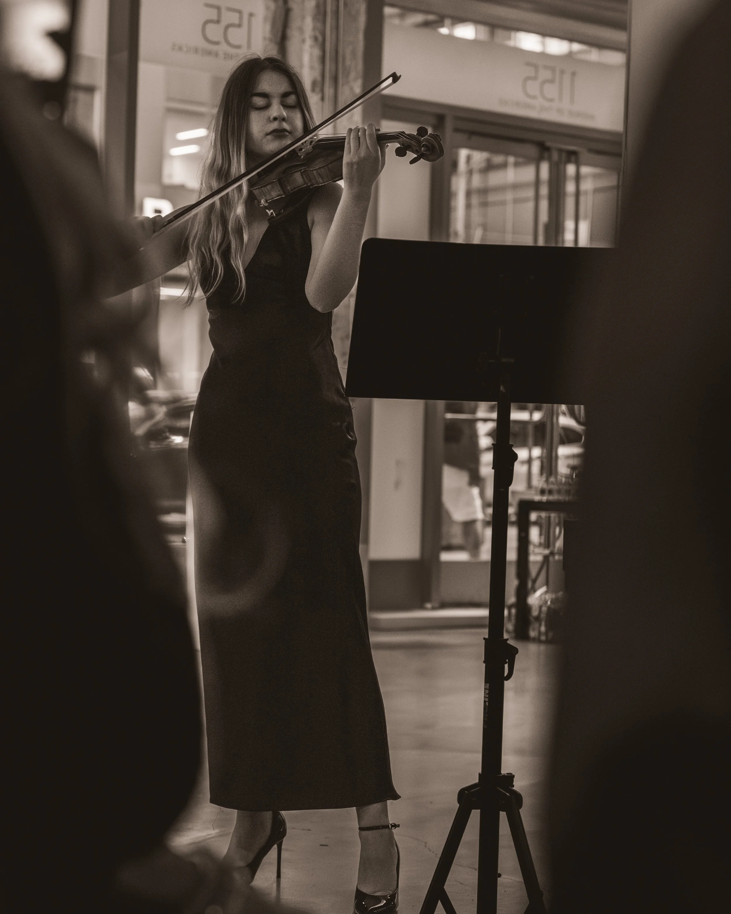 A woman in a black dress and high heels is playing the violin, standing in front of a music stand, with her eyes closed, in an indoor setting.