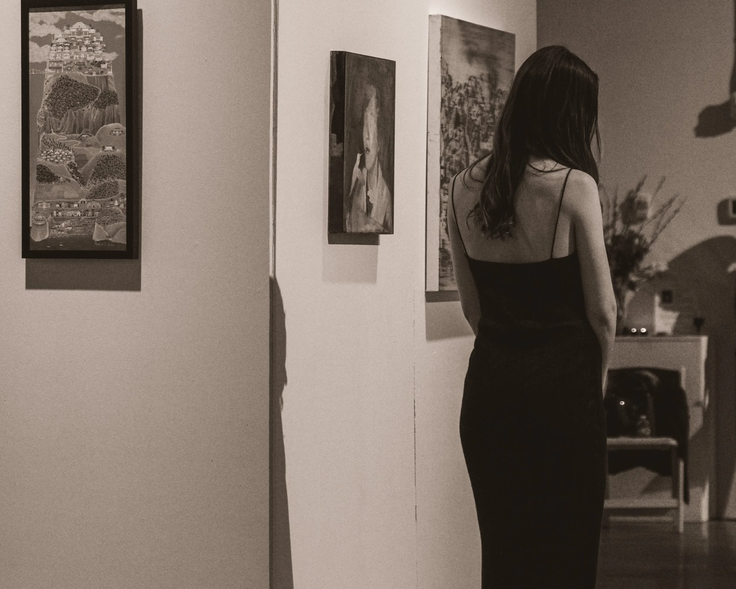 A woman with long dark hair wearing a black spaghetti strap dress is at an art gallery, looking at paintings on the wall.