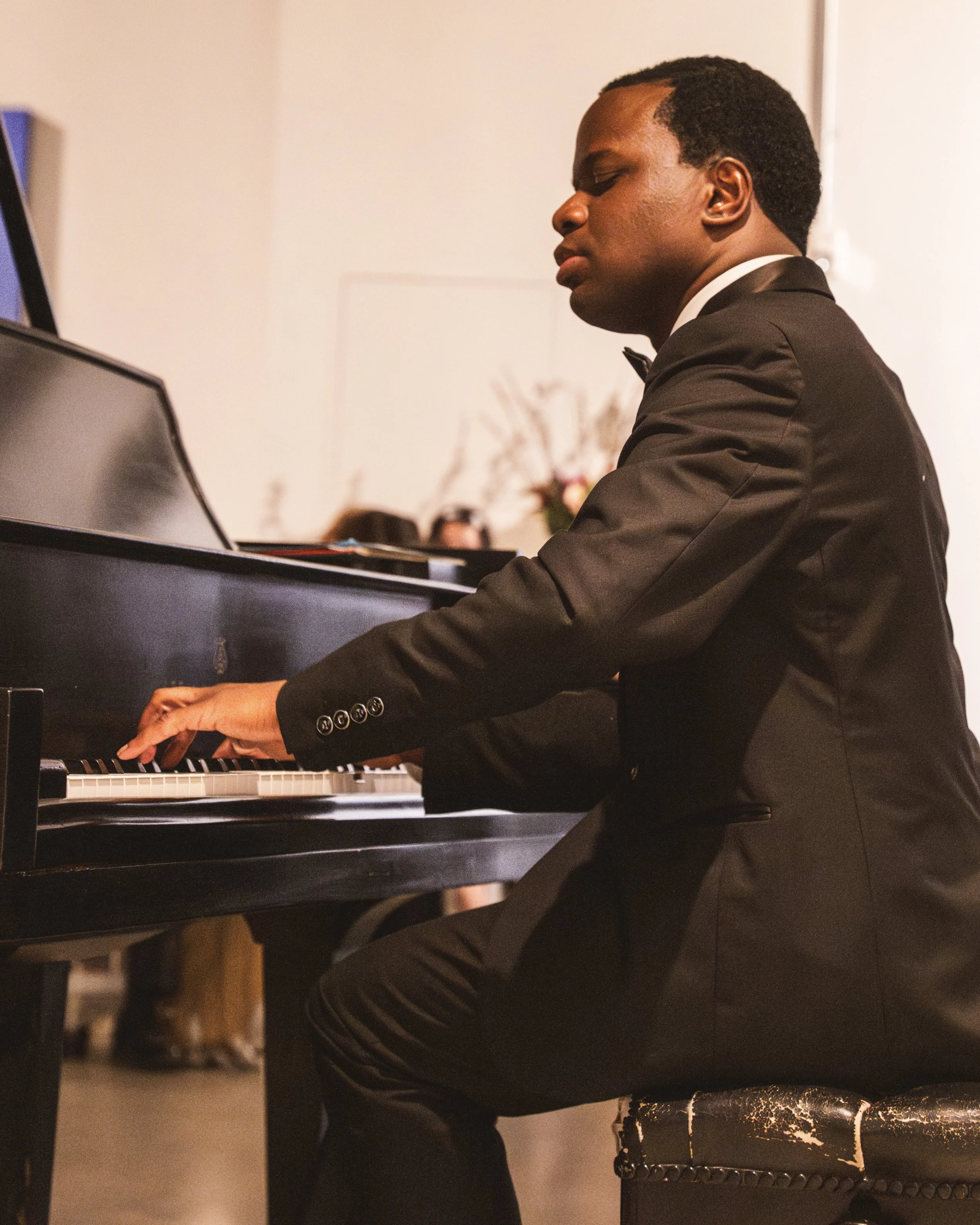 A man dressed in a black tuxedo playing a grand piano, with a serious expression and eyes closed.