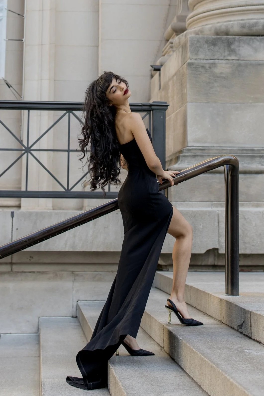 A woman with long, curly black hair and red lipstick wearing a strapless black gown and high heels poses on stairs with a metal railing outside a building with stone walls.