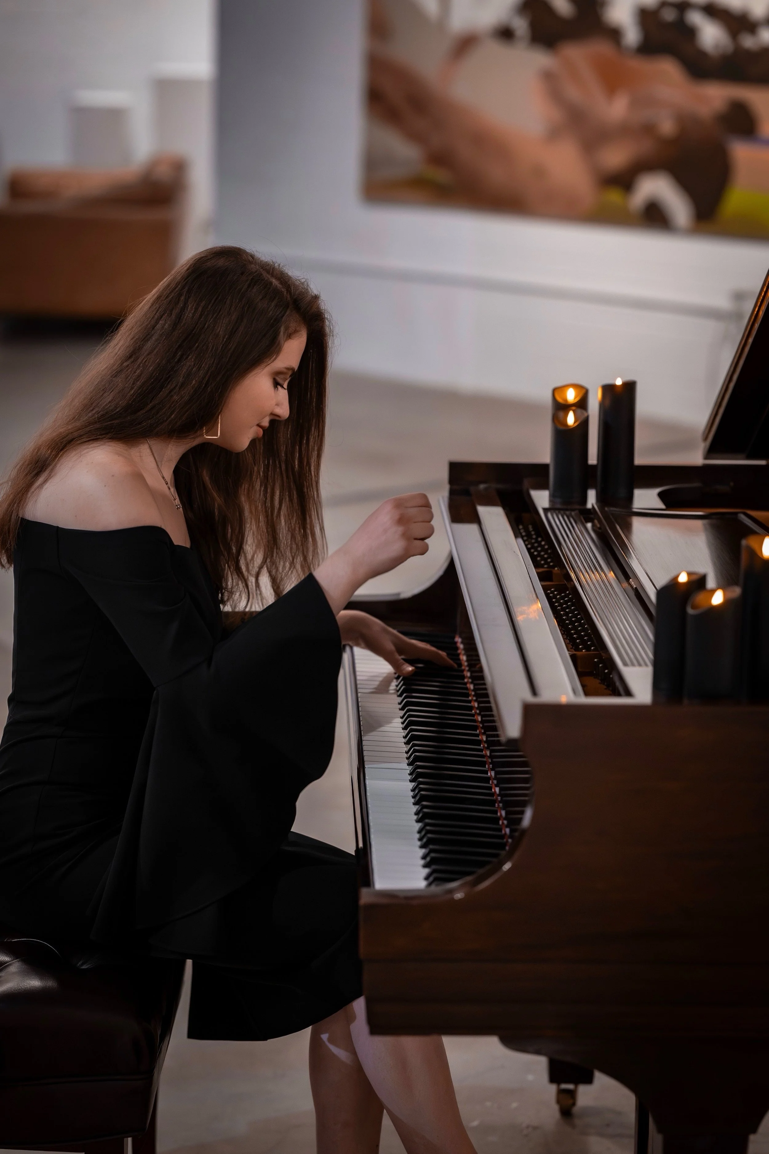 A woman with long brown hair in a black off-shoulder dress playing a grand piano in a room with soft lighting.