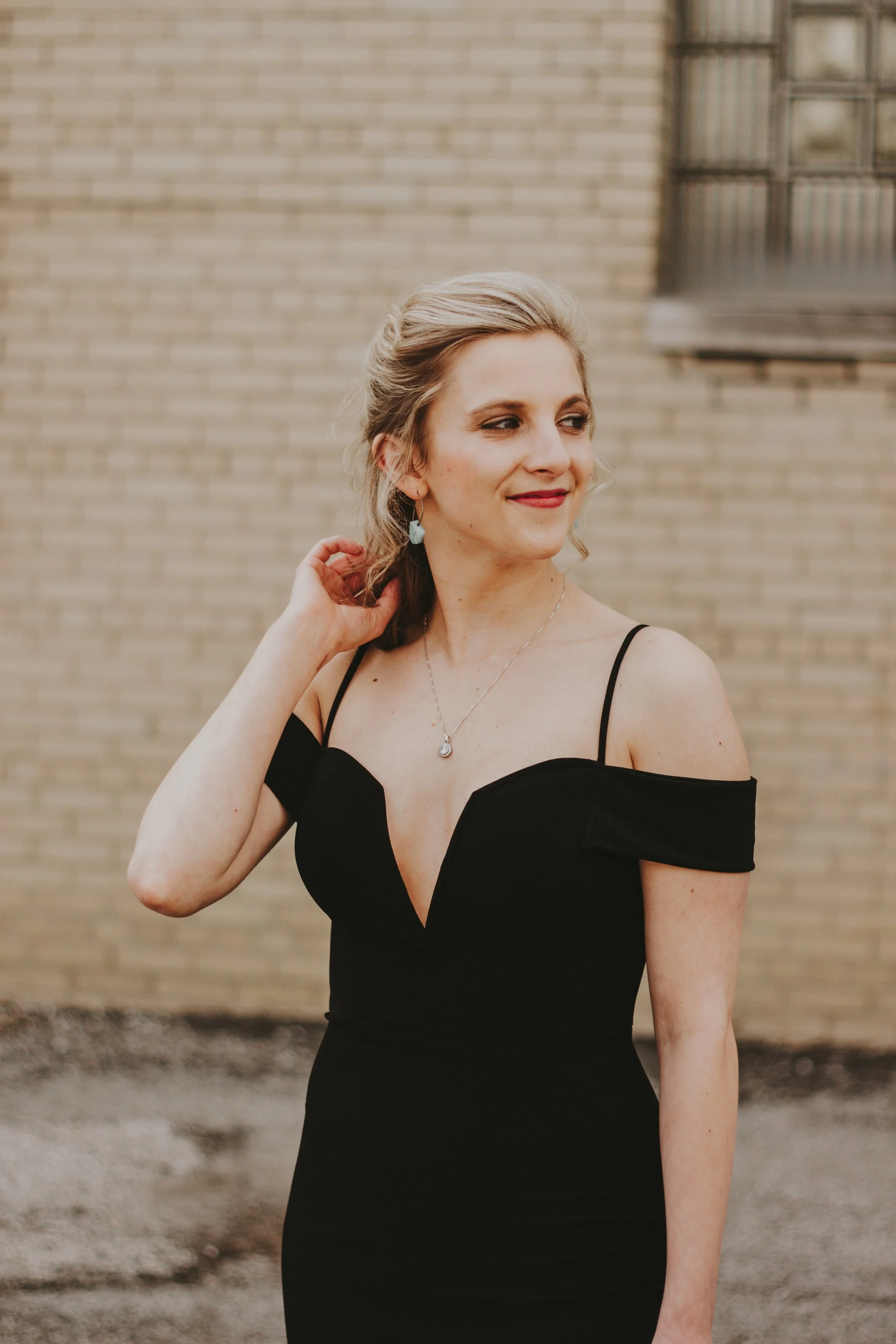 A woman in a black off-shoulder dress standing outdoors against a brick wall, touching her hair and looking to the side.