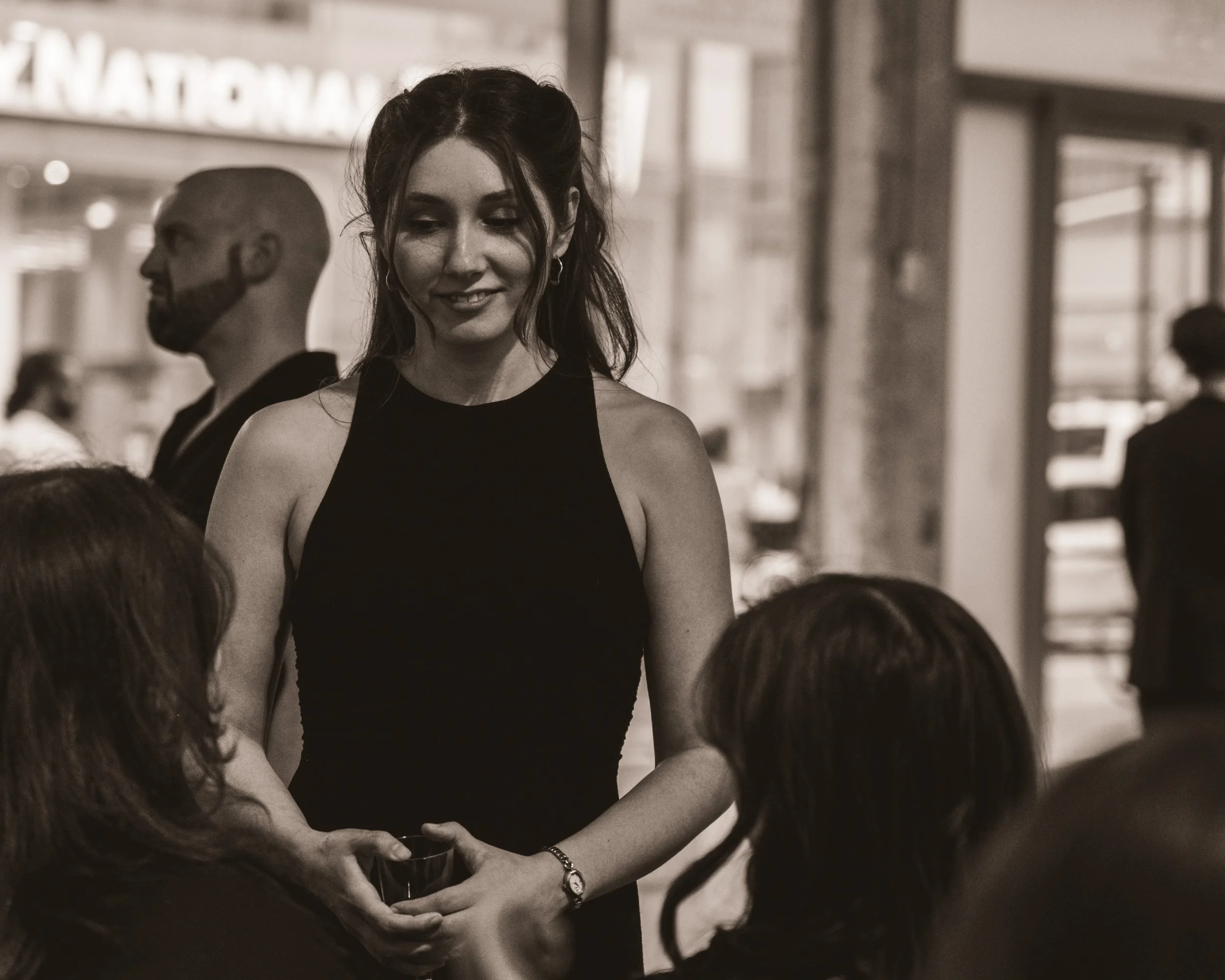 A woman in a black sleeveless dress smiling while serving drinks to two women seated at a table in a crowded indoor setting.