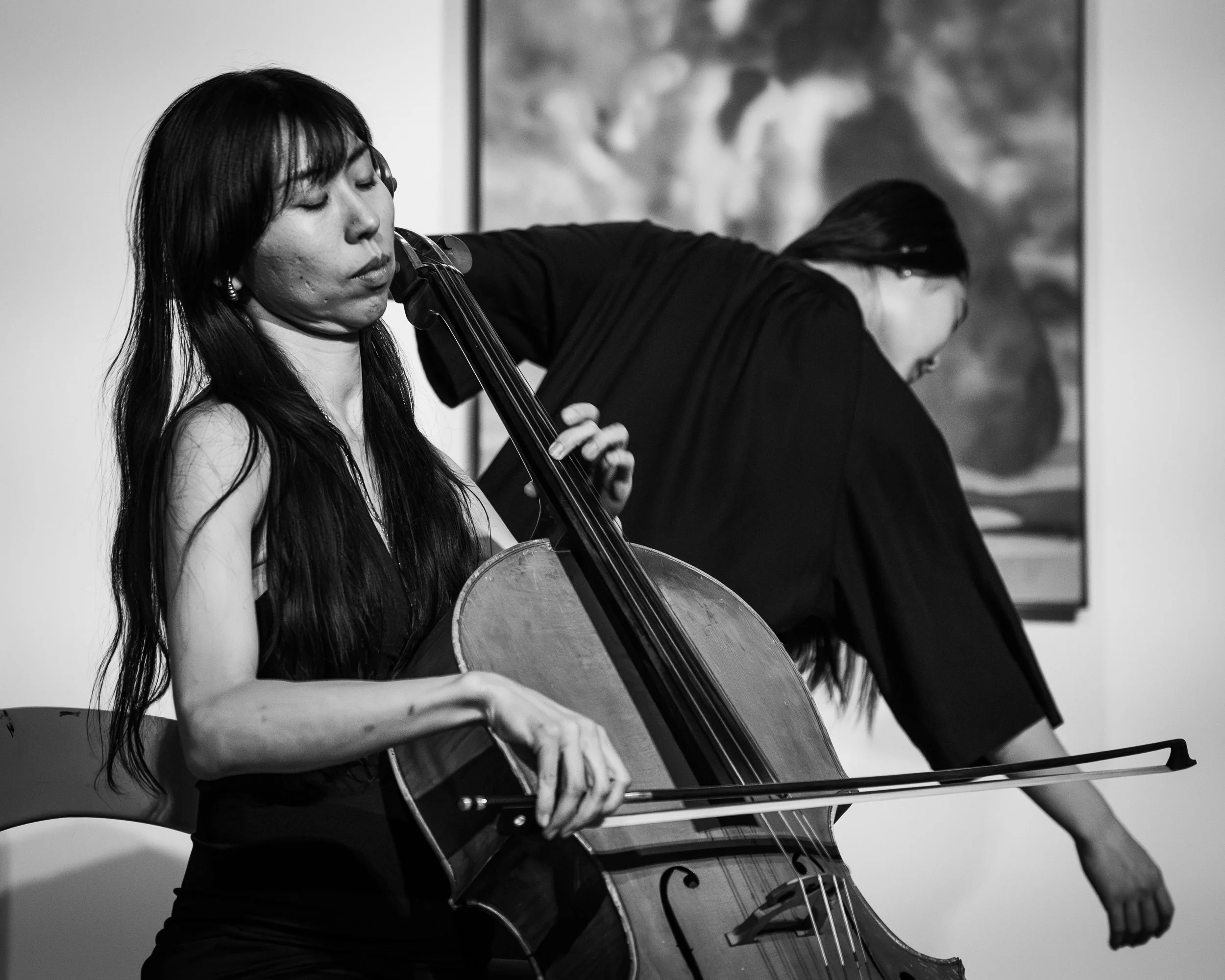 A woman playing a cello during a performance, with another person in the background.