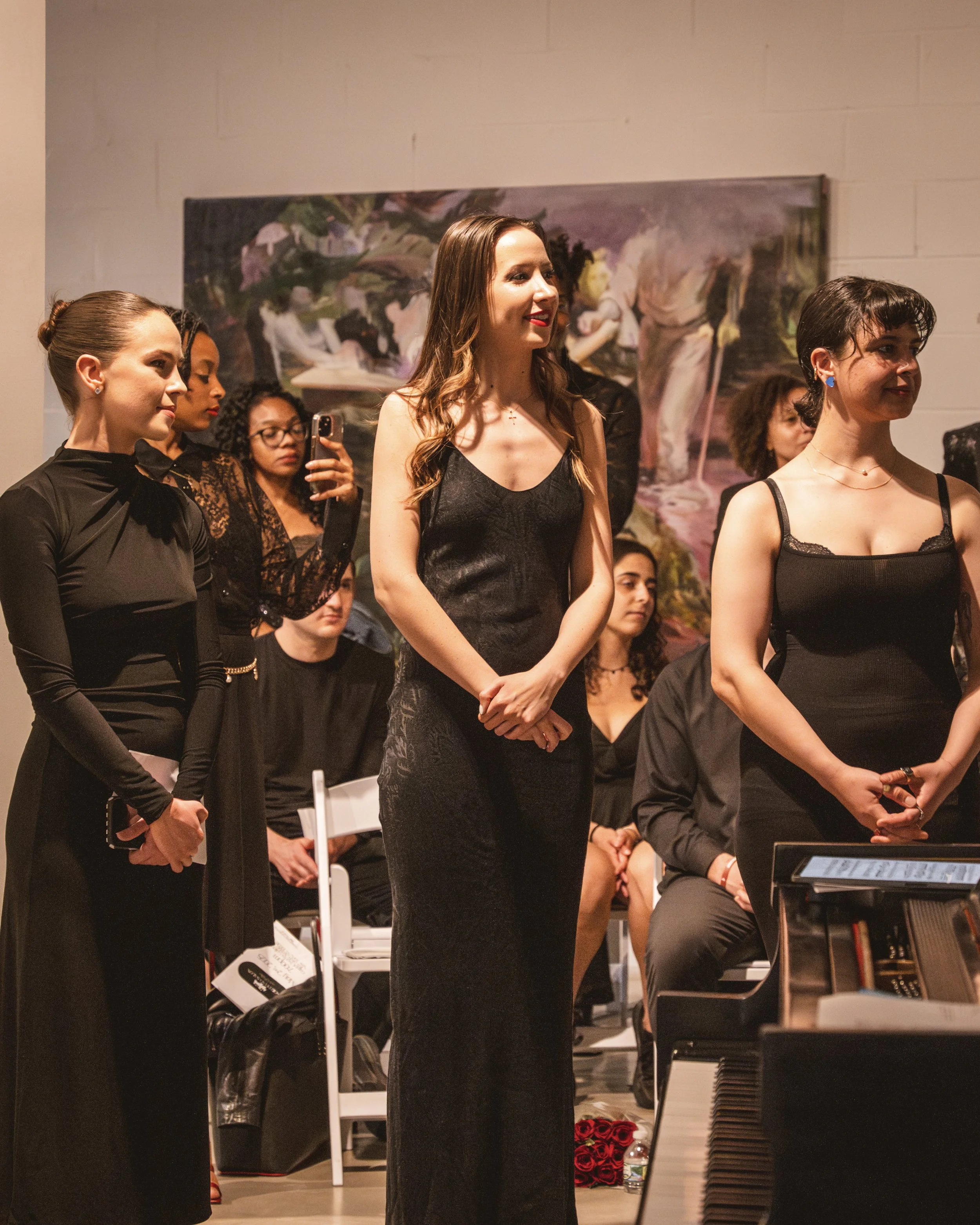 A group of women in black dresses standing in a room, with some seated in the background, next to a piano, at an indoor event or performance.