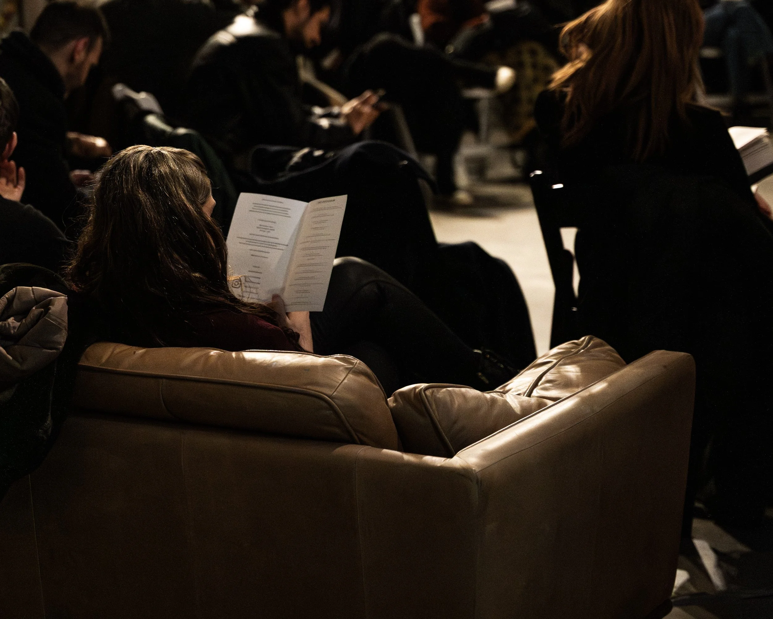 People seated in a dimly lit room, reading from papers or programs, with some looking at their phones.
