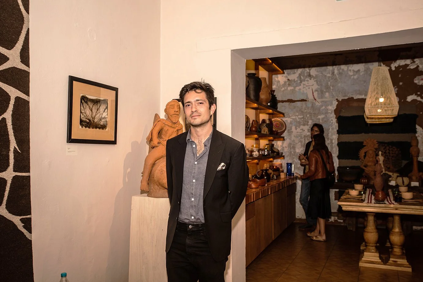 Man in black suit standing in art gallery with people shopping in background.