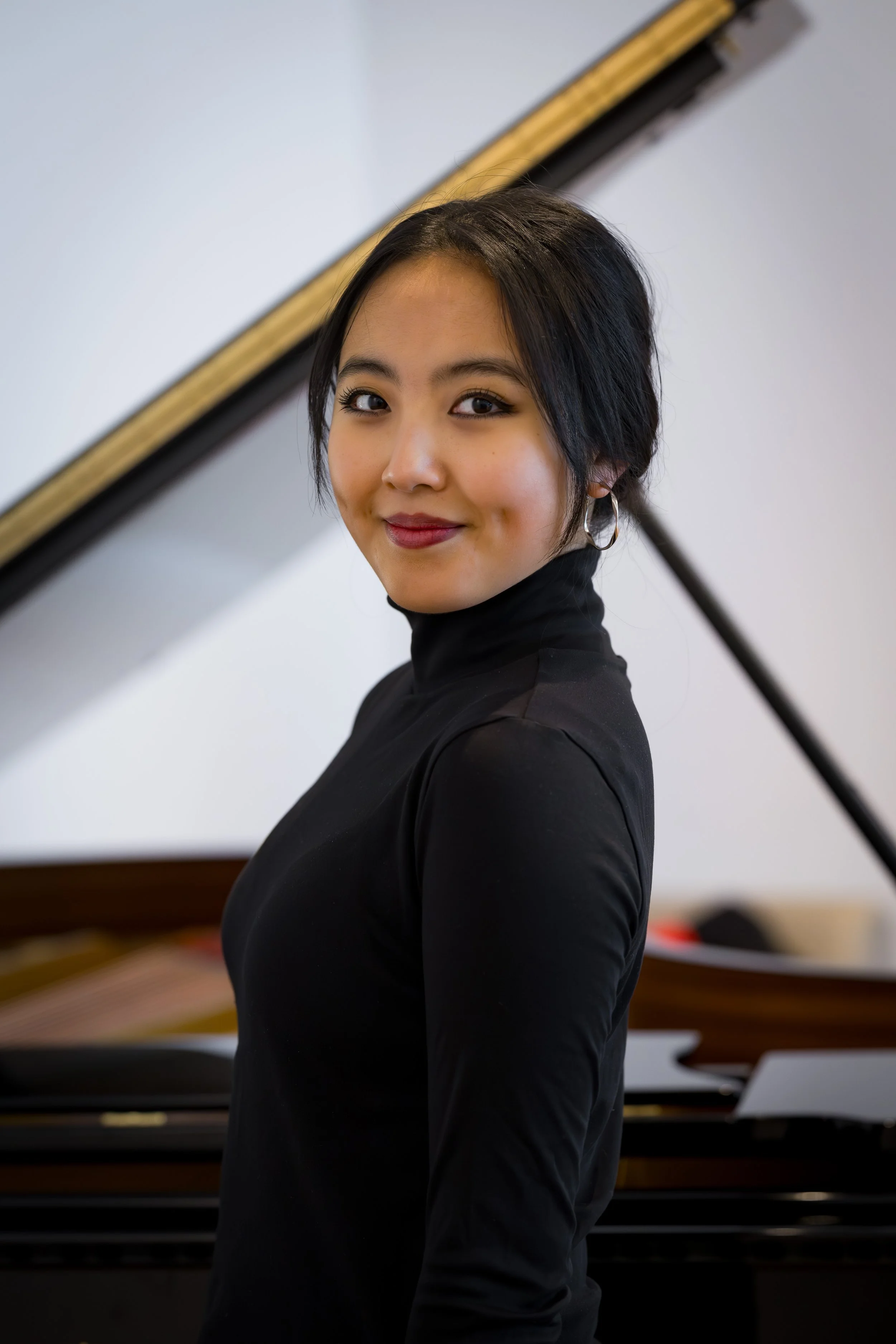 A young woman with dark hair, wearing a black turtleneck, smiling, standing in front of a grand piano.