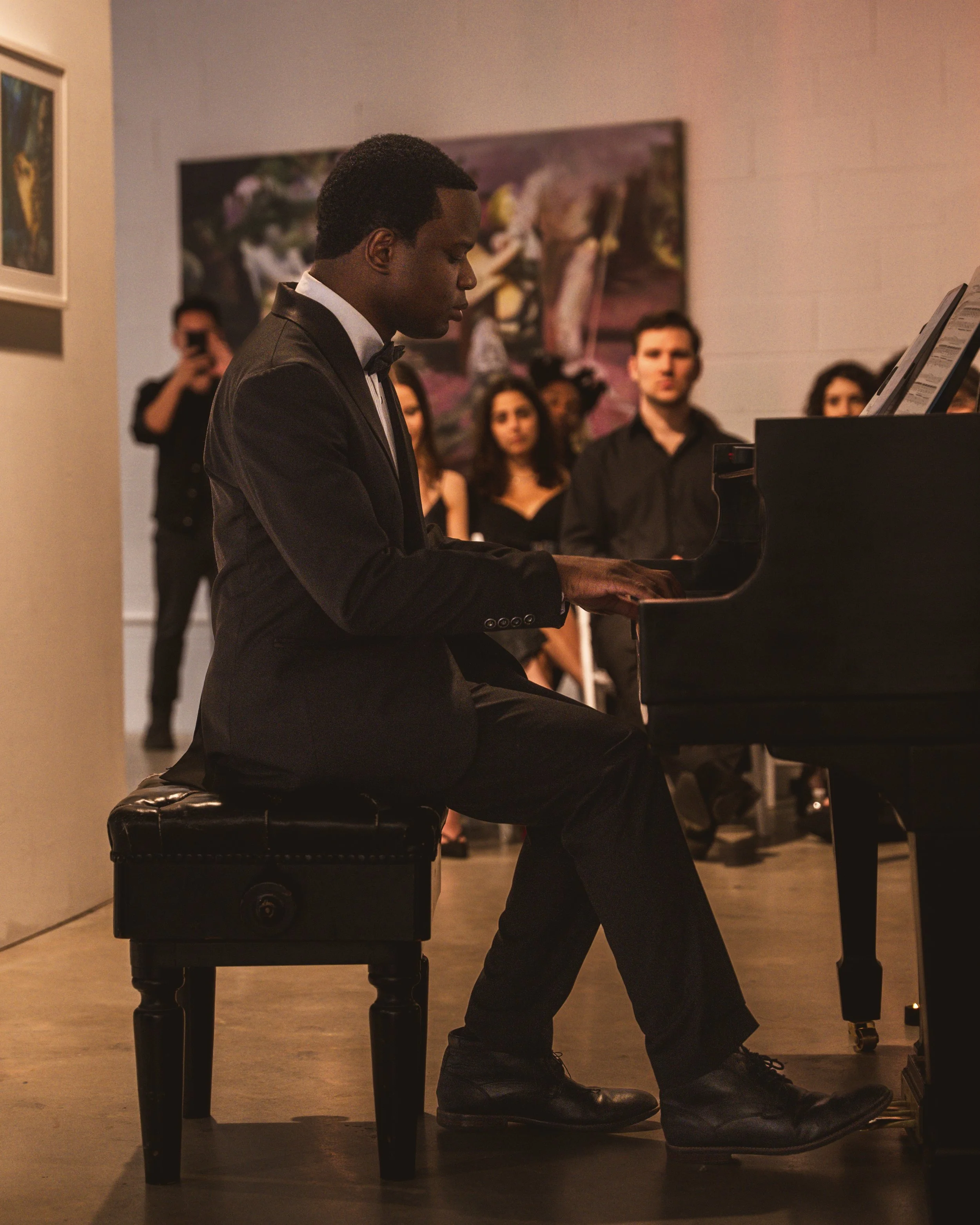 A man in a tuxedo playing the piano at an art gallery, with an audience watching.