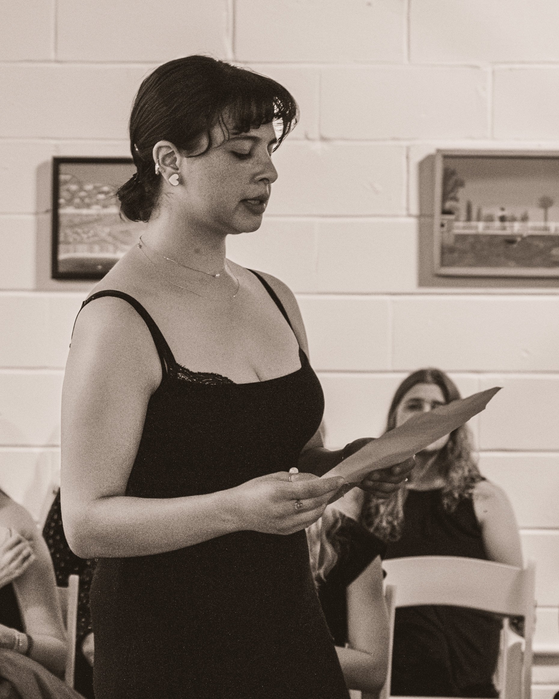 Woman in a black tank top reading from paper at indoor event with seated audience