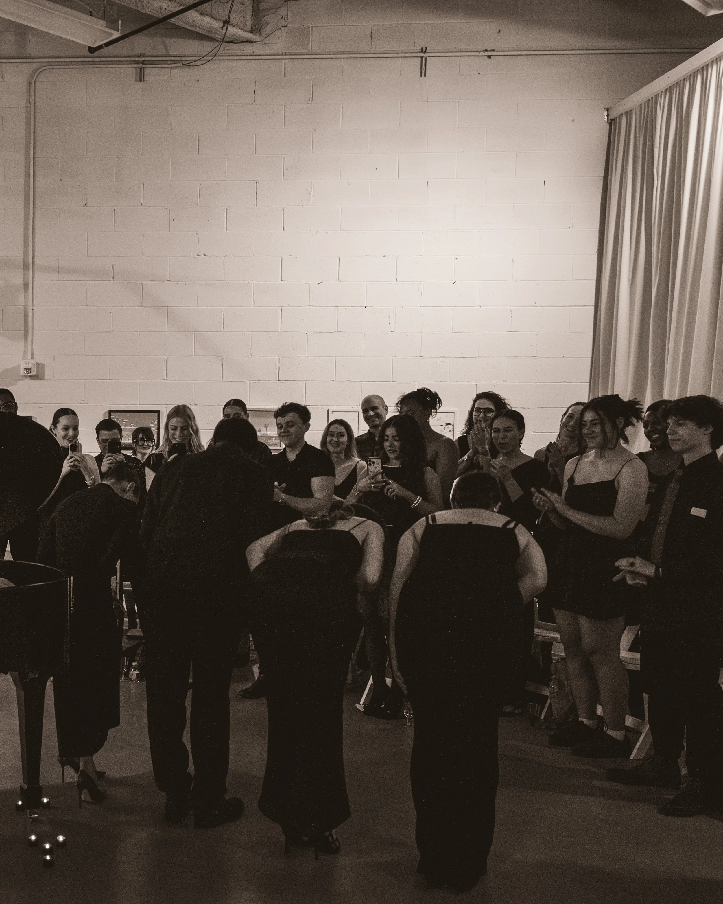 A group of people at an event, some taking photos, standing in front of a plain wall with framed pictures, with a curtain on the right side.