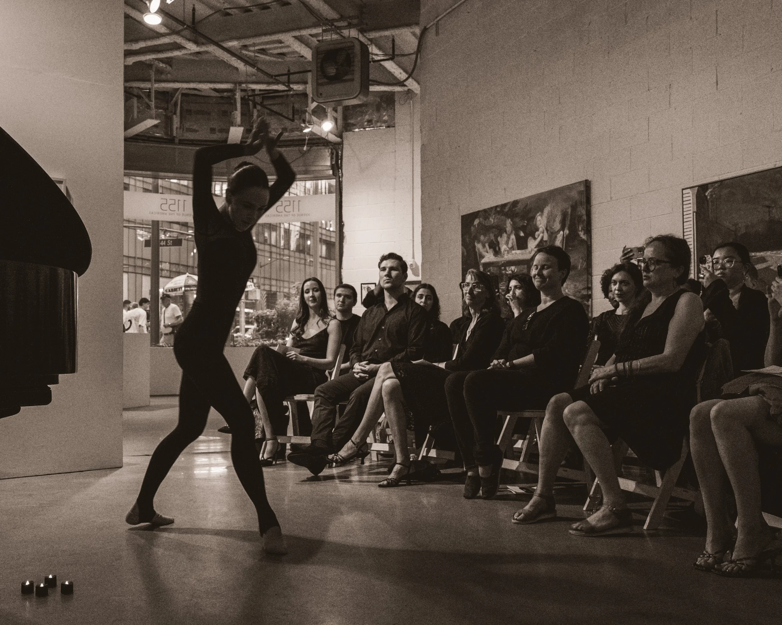 A woman performing a dance or ballet in front of an audience seated indoors in an art gallery or studio. The audience appears engaged, with some taking photos or videos. The scene is in black and white.