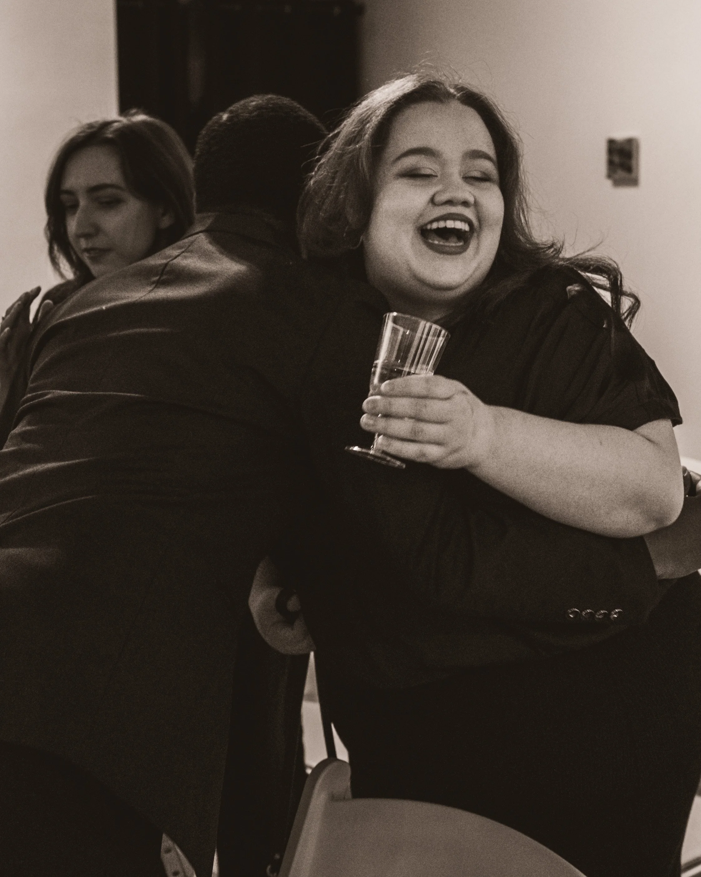Two women and a man hugging, the woman in front smiling and holding a glass, in a celebratory setting.