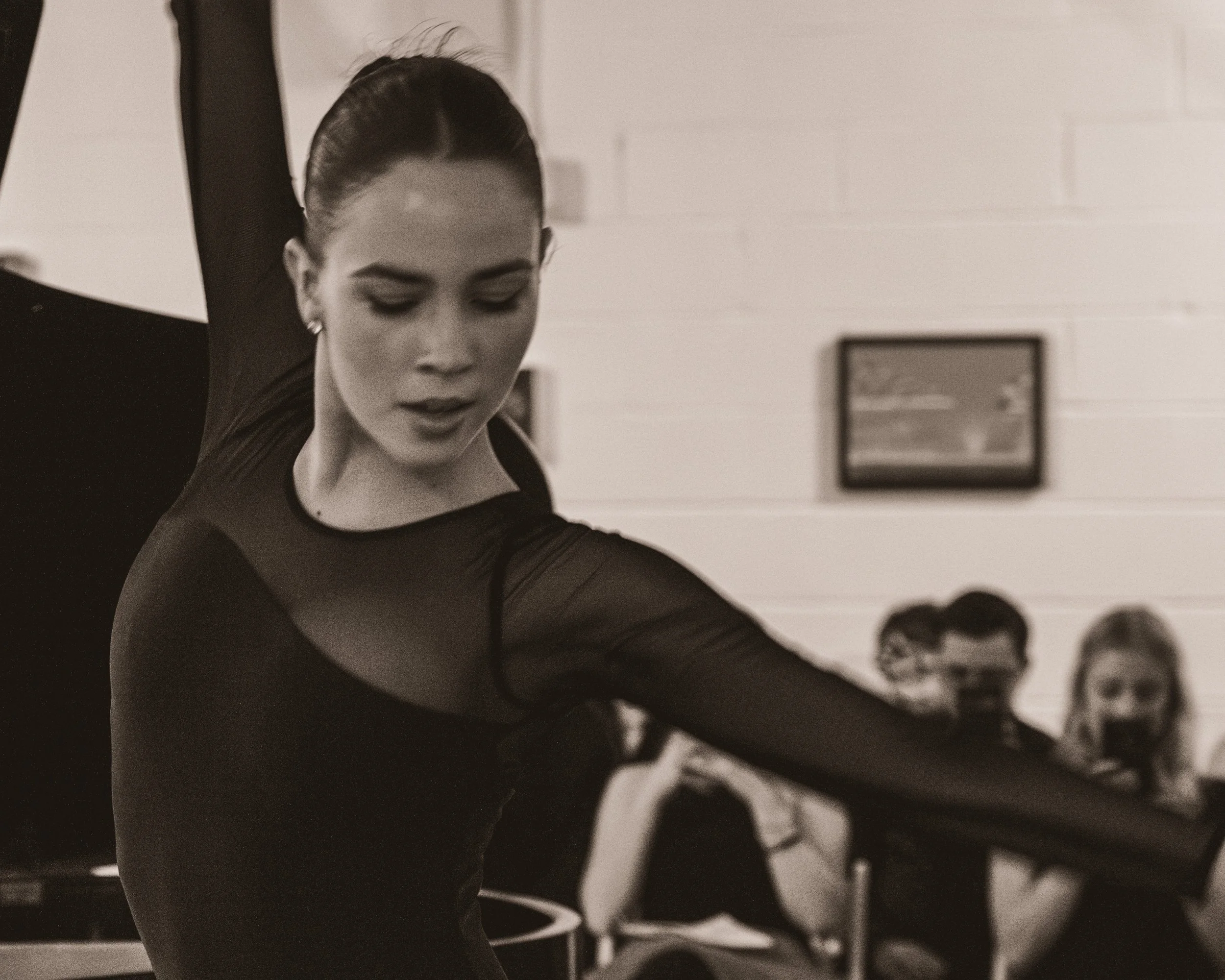 A woman performing a ballet dance, wearing a black leotard with sheer sleeves, in a dance studio with a white brick wall background, while others are sitting and watching in the background.