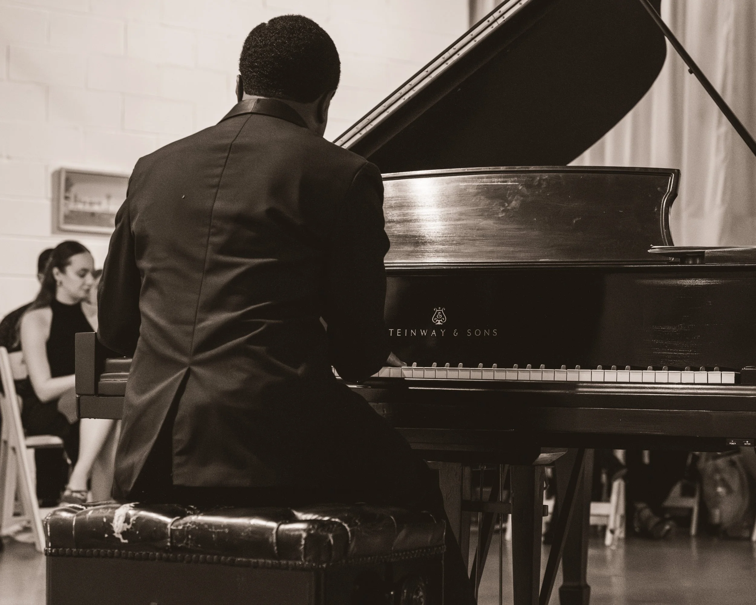 A man in a dark suit playing a grand piano, with a woman in a black dress sitting in the background.