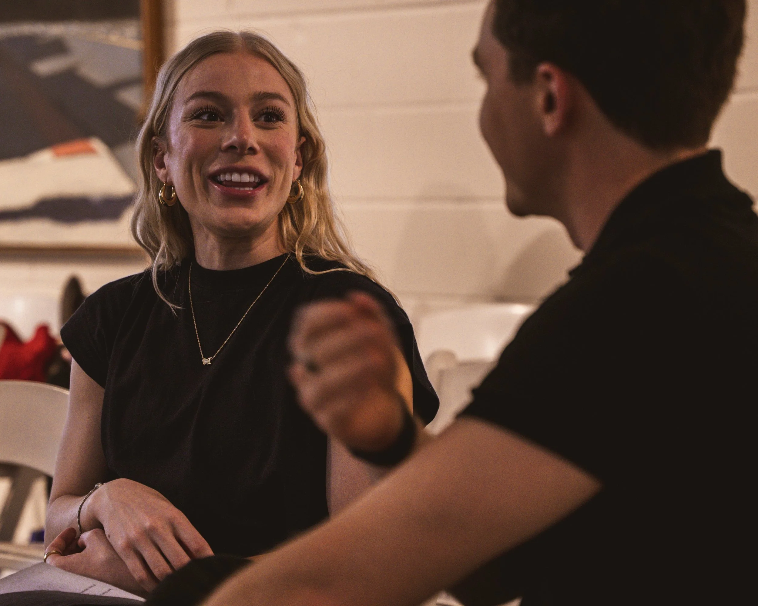 A woman with blonde hair, wearing a black top, gold hoop earrings, and a gold necklace, smiling and talking to a man with short brown hair, wearing a black shirt. They are sitting indoors, engaging in conversation.
