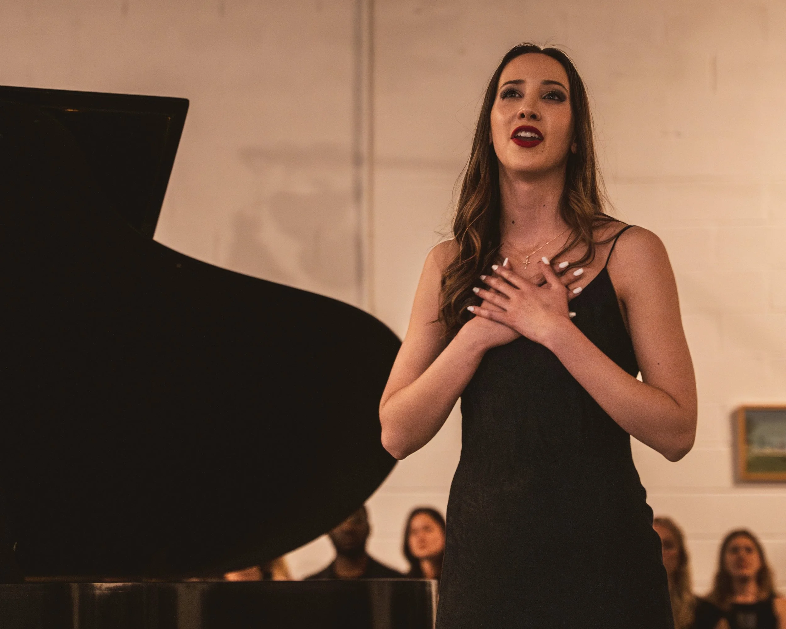 A woman in a black dress singing passionately at a piano with her hands on her chest, with an audience in the background.
