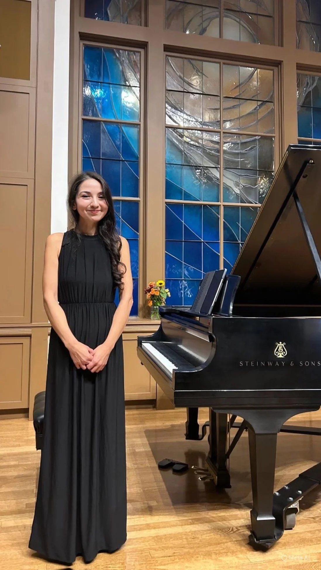 A woman in a long black dress standing next to a grand piano inside a room with stained glass windows and wooden paneling.