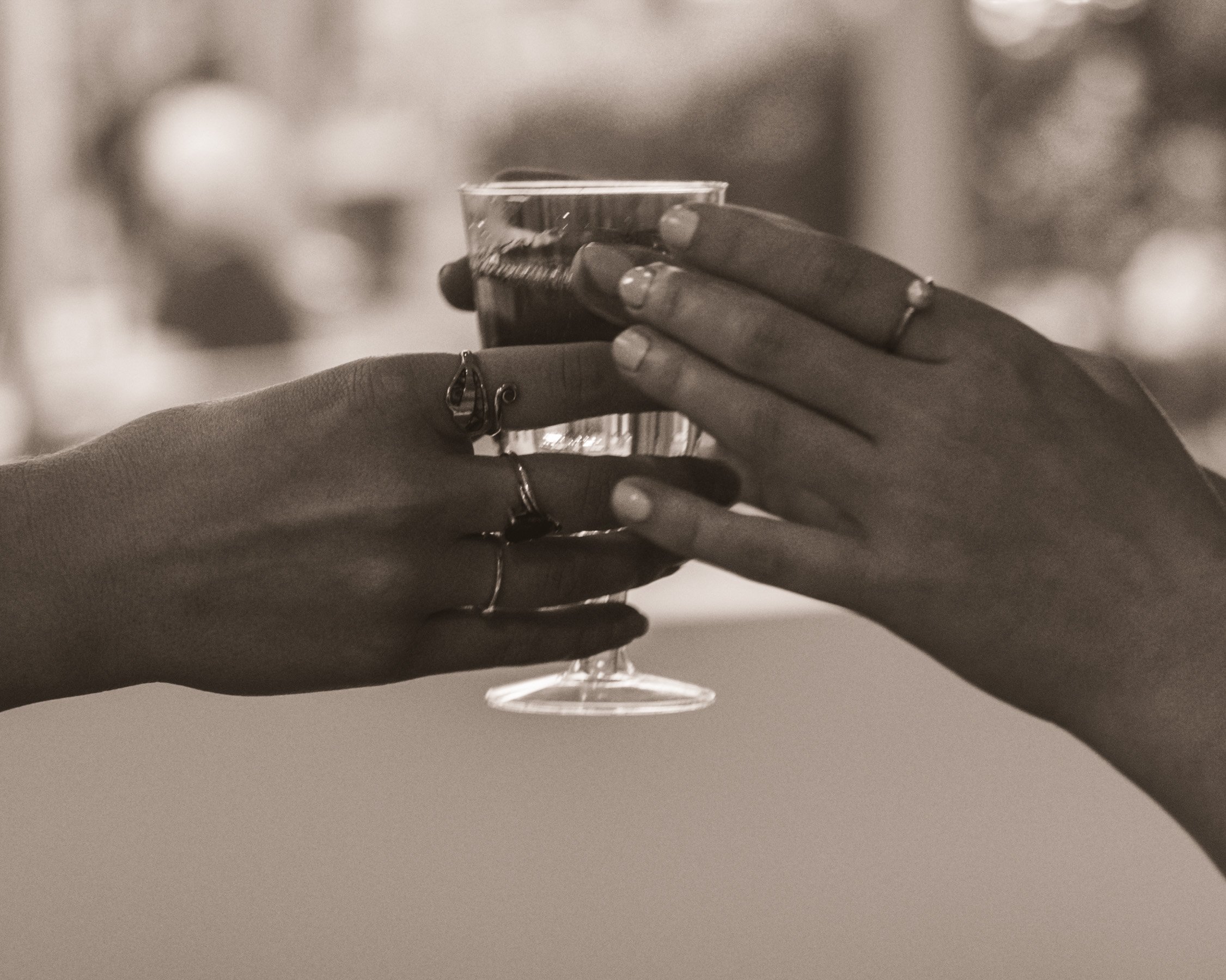 Two hands, adorned with rings, holding a glass with a dark beverage, set against a blurred background.