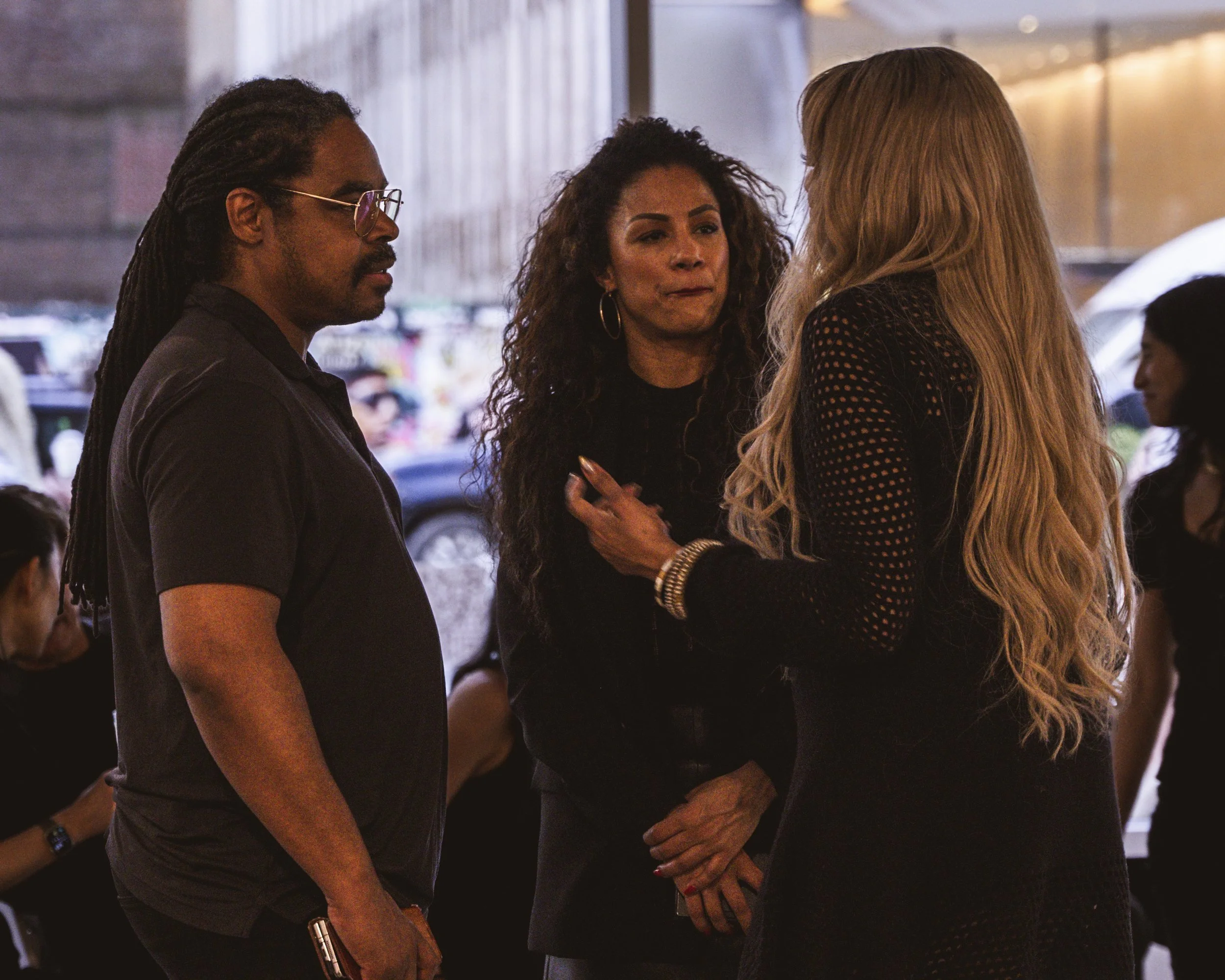 Three women in conversation indoors, with a blurred background of people and window display.
