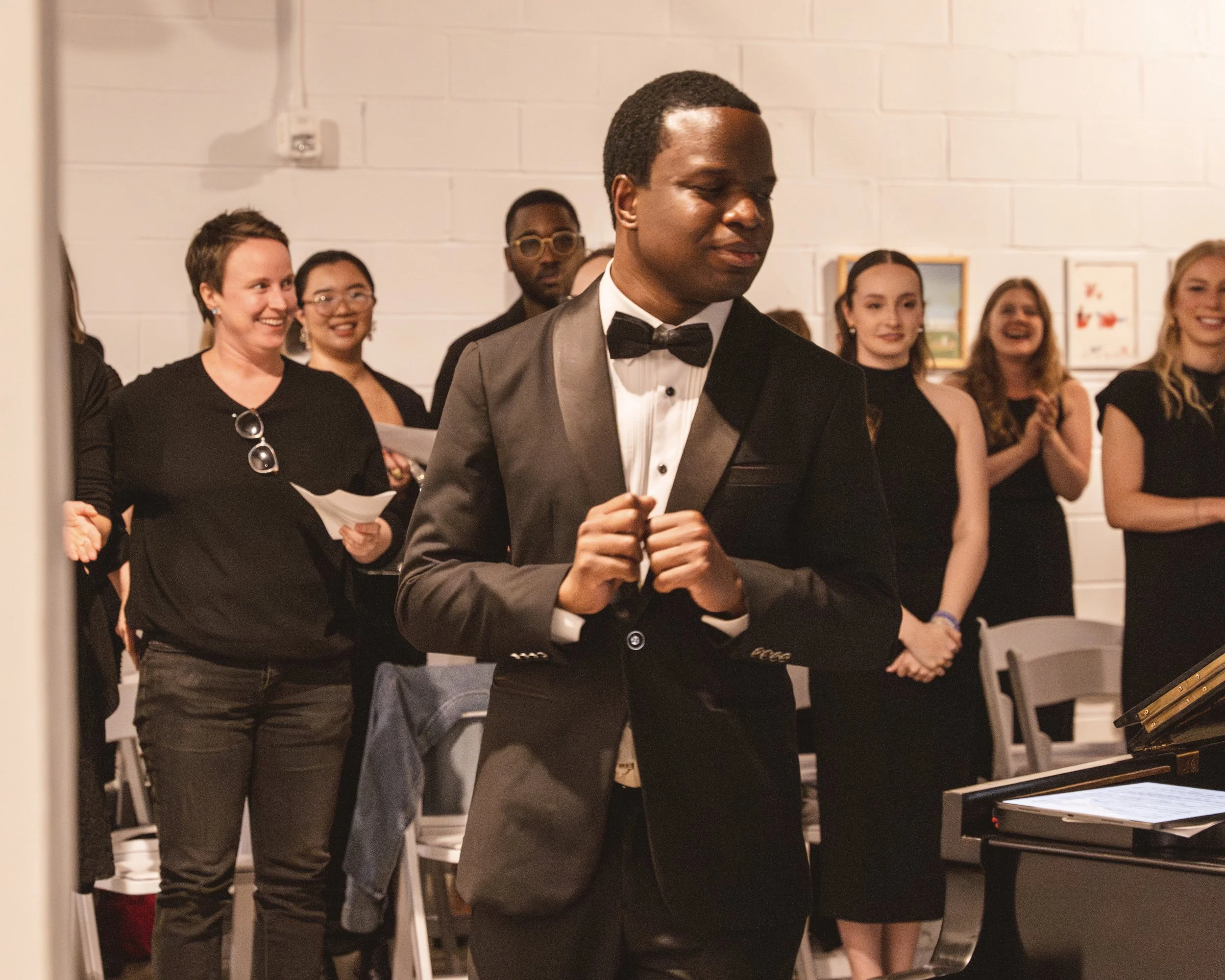 A young man in a tuxedo stands in front of a group of people, possibly at a concert or event. The people behind him are clapping and smiling, dressed in black. There is a piano in the foreground.
