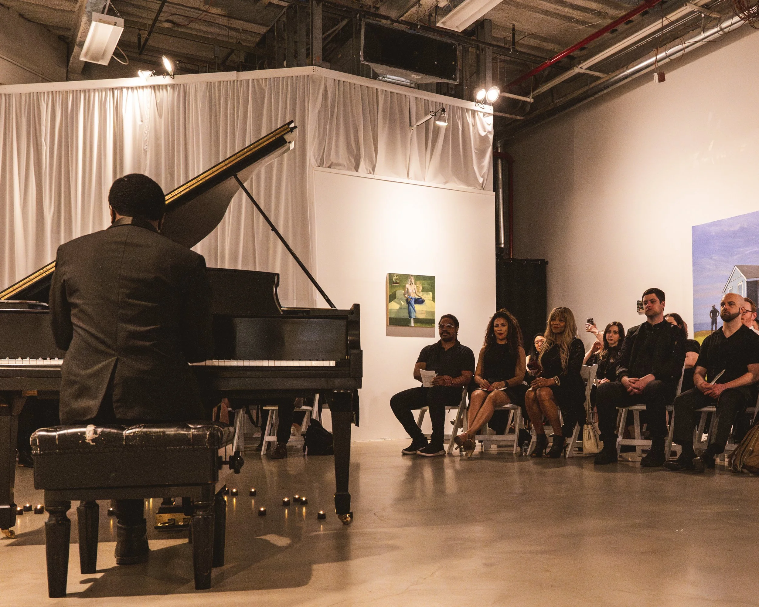 A pianist performs on a black grand piano in front of an audience in an art gallery or event space. The audience is seated and watching, with some individuals taking photos or notes, and artworks are displayed on the white walls.