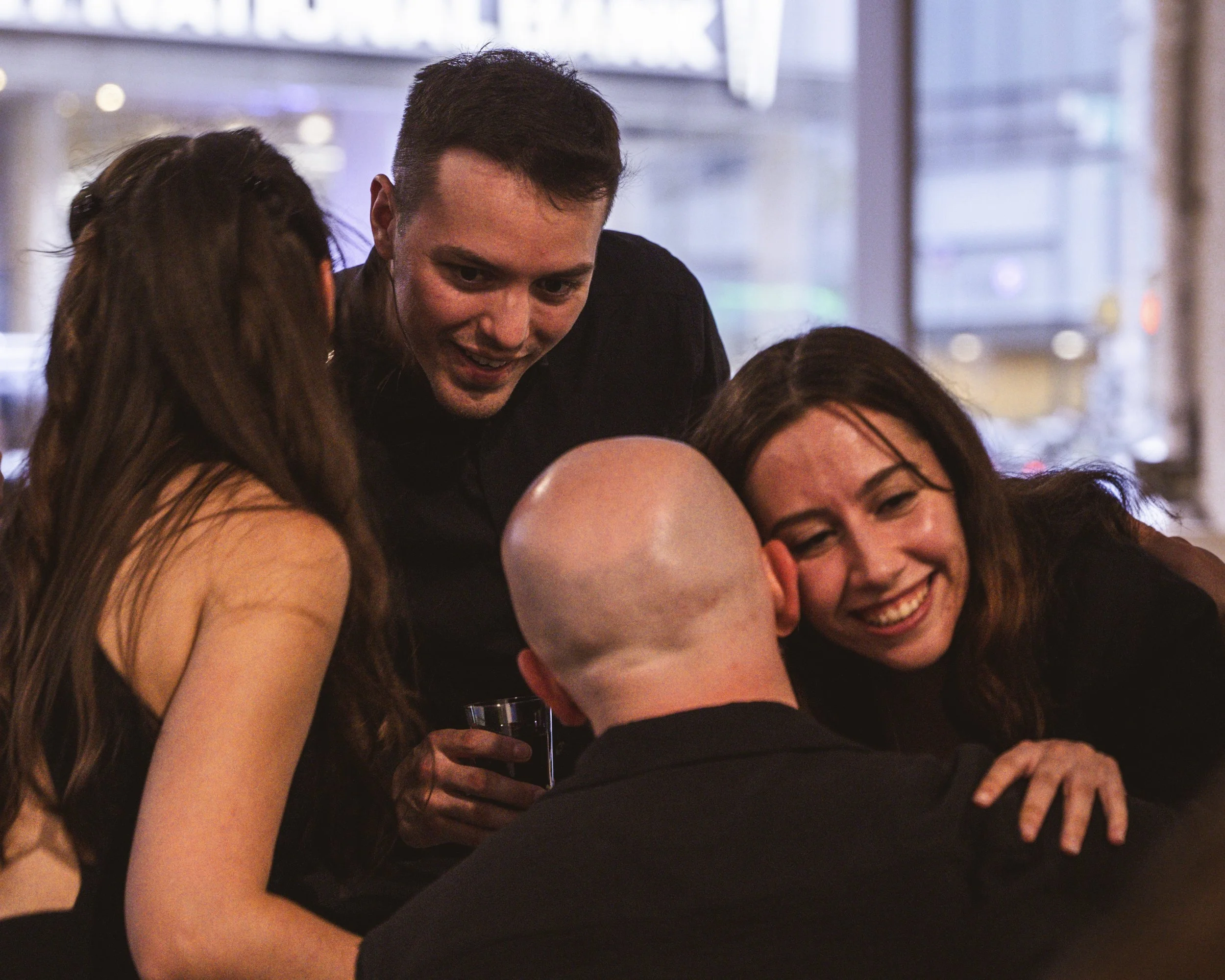 Four friends are gathered close together, smiling and laughing, sharing a joyful moment in a cozy indoor setting.