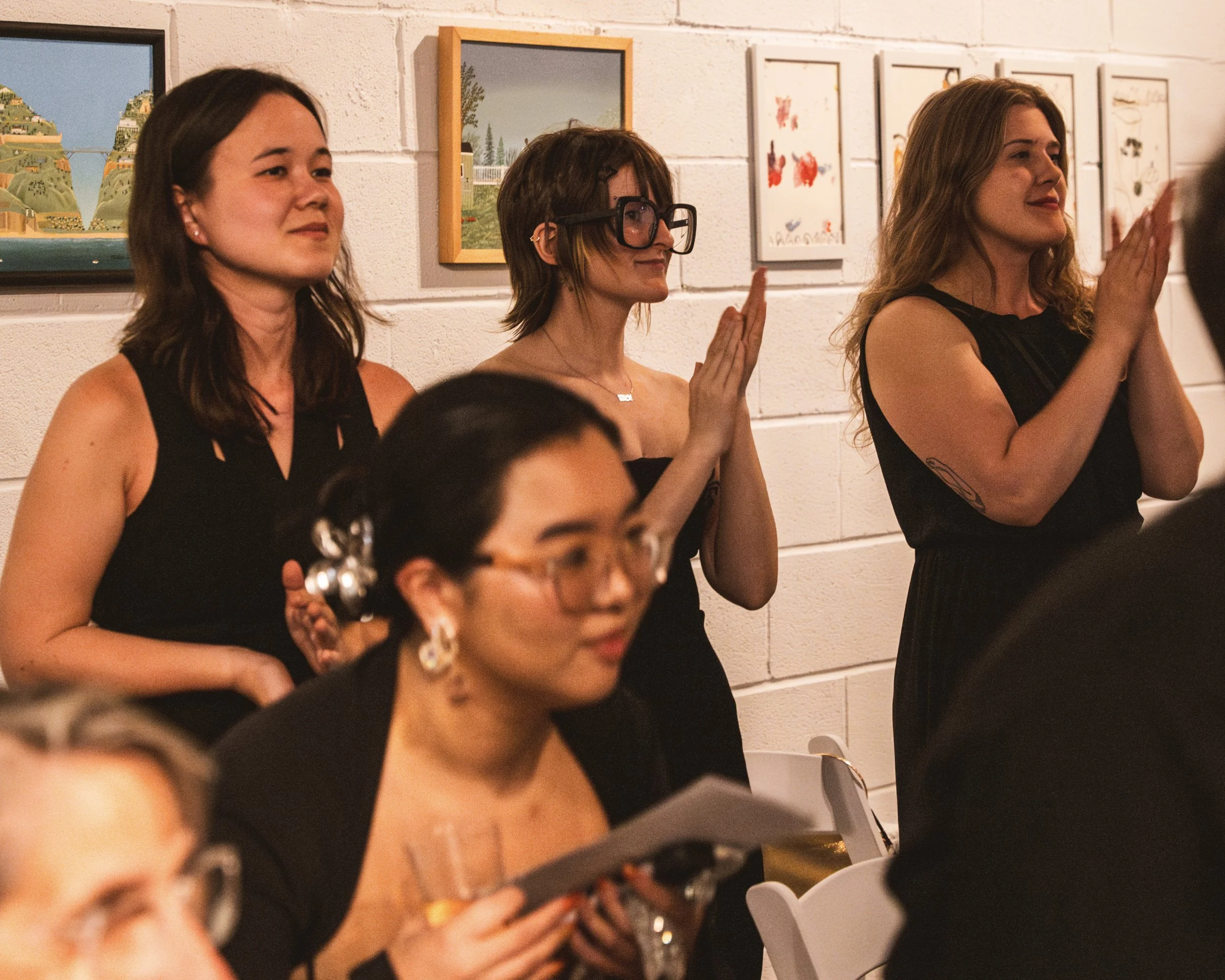 Three women standing against a white wall with framed artwork, clapping and attentively watching an event, with one woman in the foreground holding a glass and a booklet.