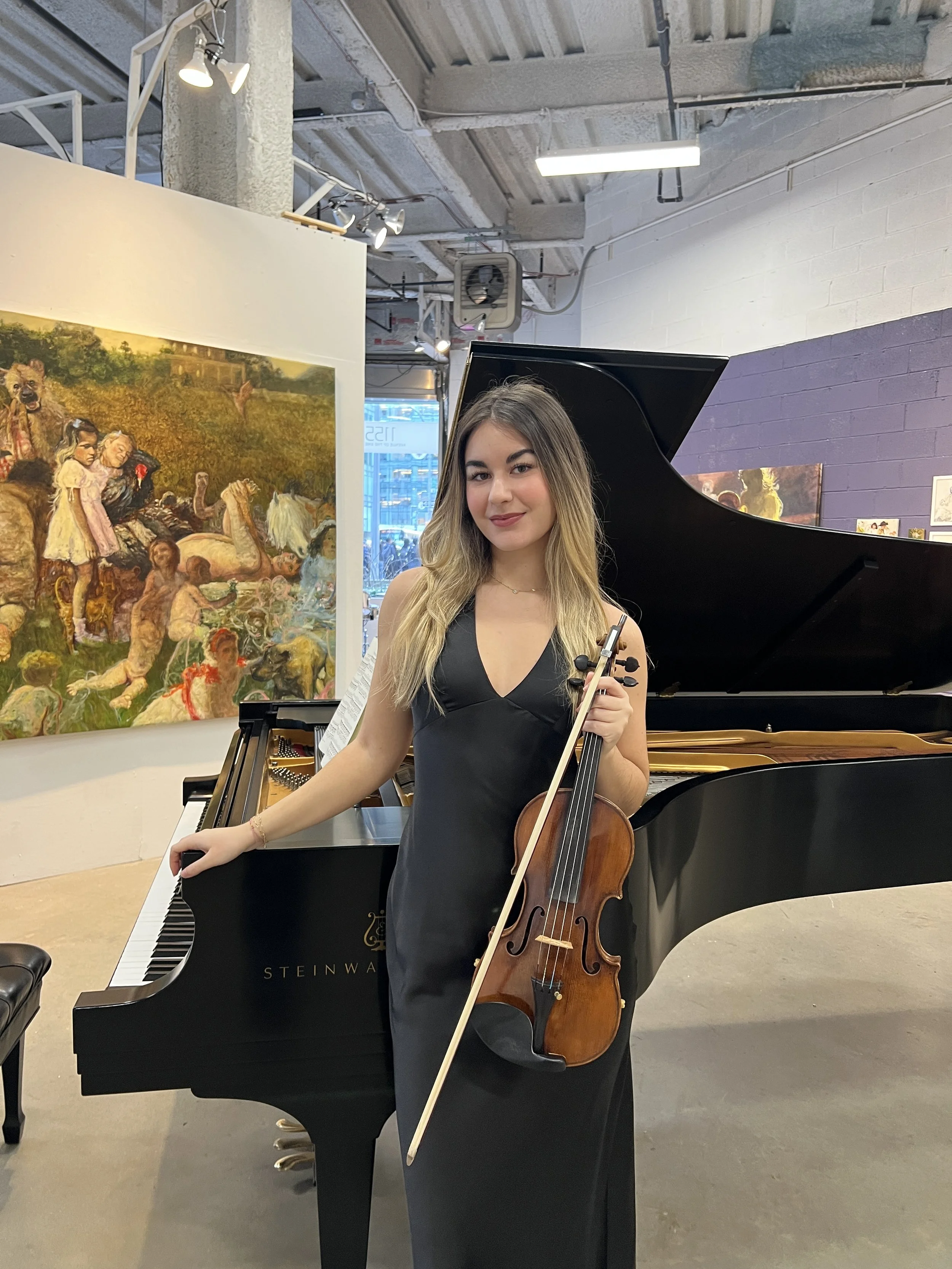 Young woman in a black dress holding a violin and bow, standing next to a grand piano inside an art gallery or music venue.