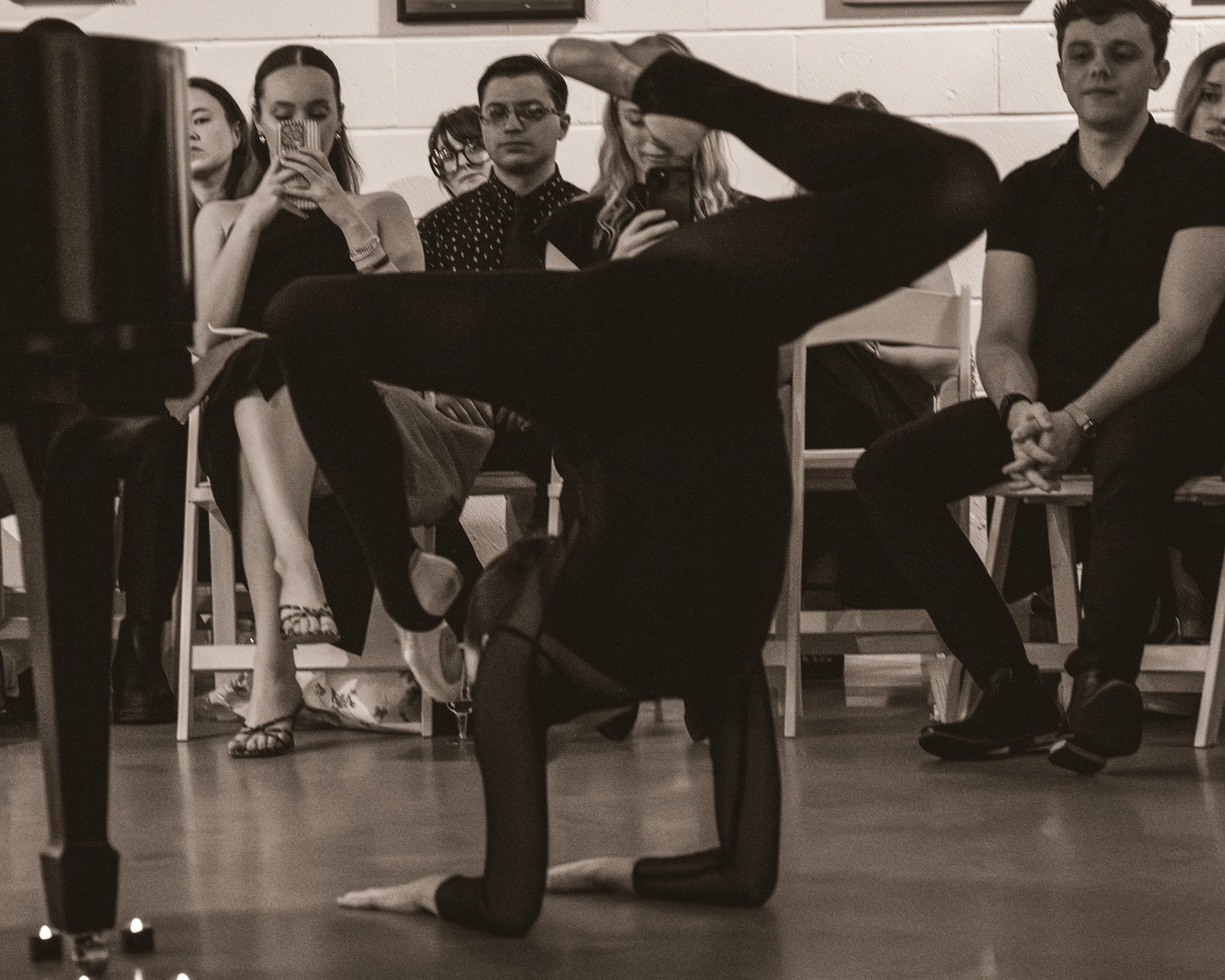 A person in black clothing performs a head-stand dance move called "handstand" in front of an audience seated on chairs, who are watching and taking photos of the performance.