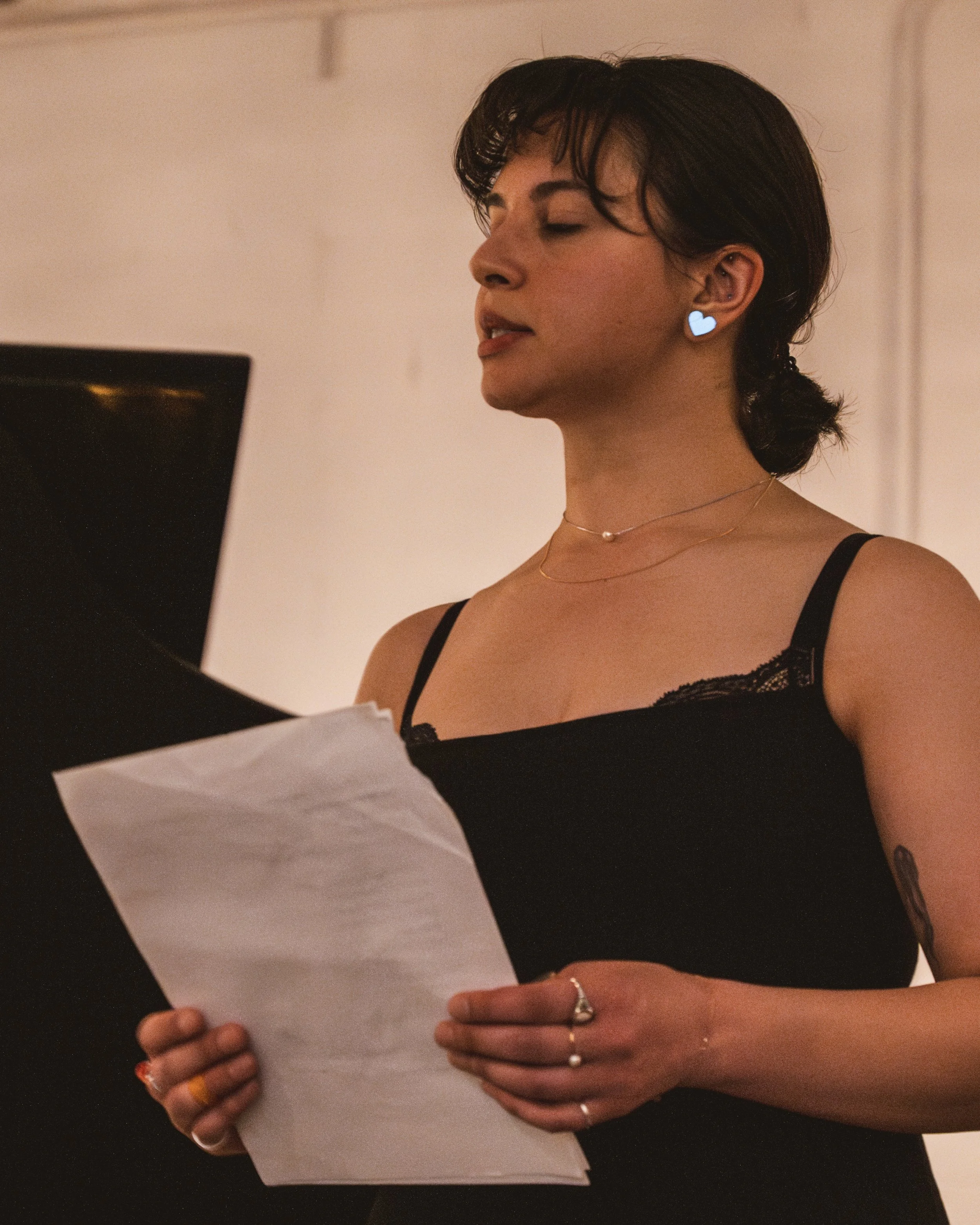 A woman with short dark hair wearing a black tank top and heart-shaped earrings is singing or speaking in front of a piano, holding a sheet of paper.