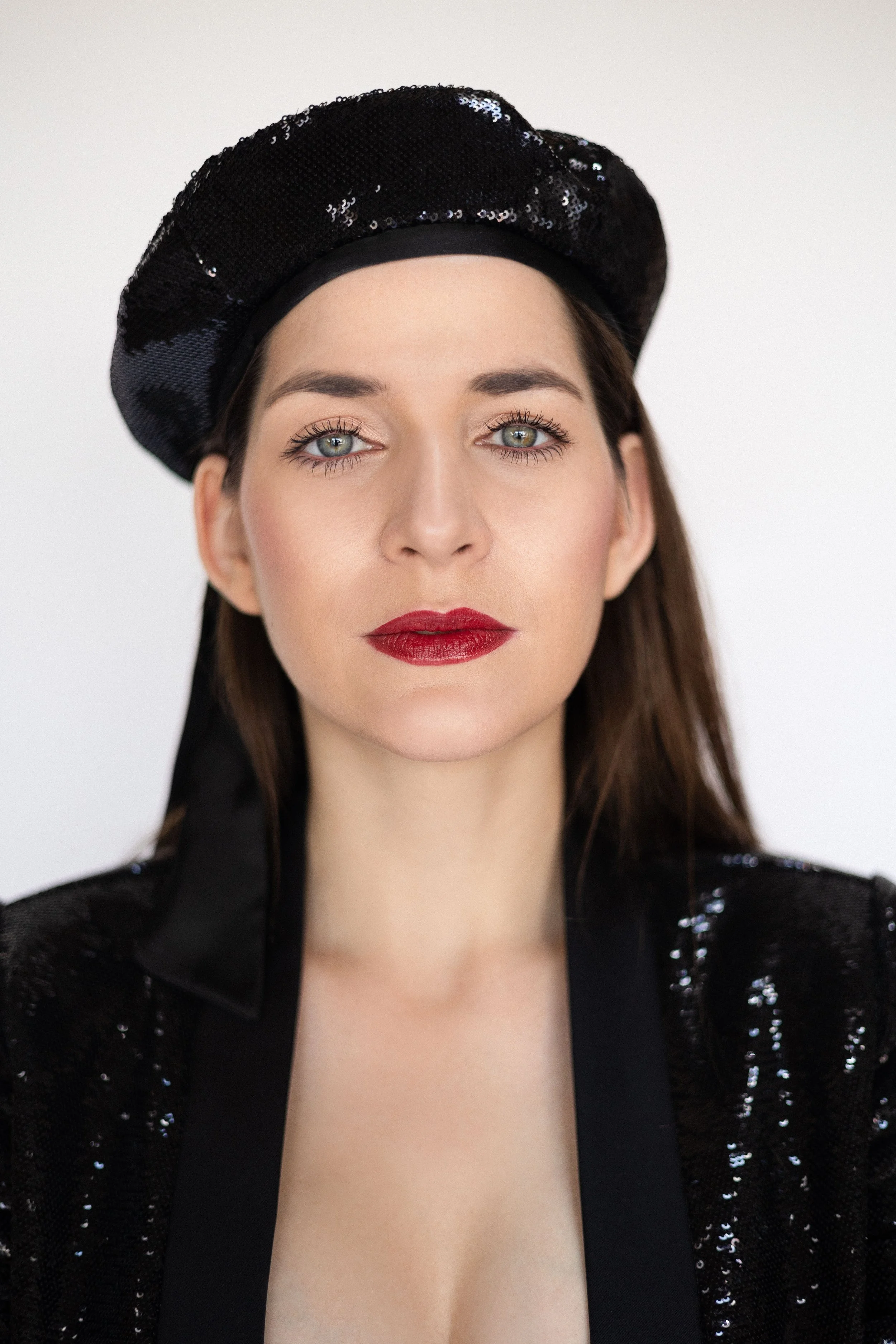 A woman with blue eyes, red lipstick, and dark brown hair wearing a black sequined beret and a black sequined jacket.