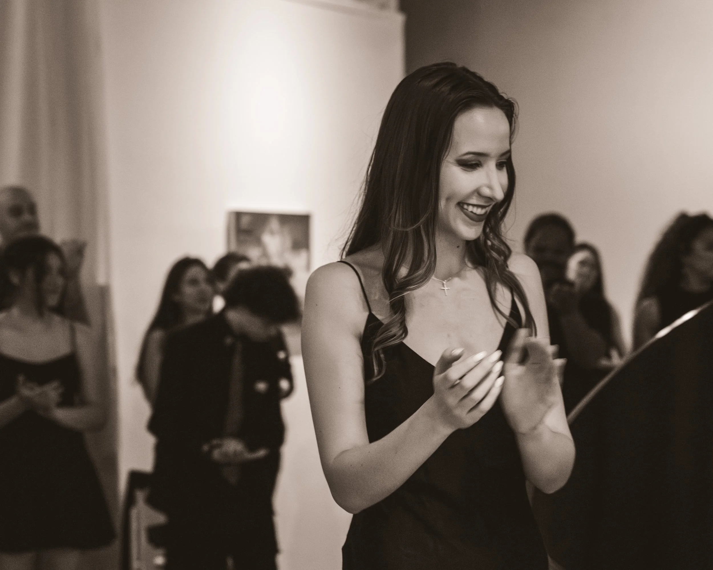 A young woman with long hair, wearing a sleeveless black dress and a cross necklace, is smiling and looking at her phone in a gallery with other women in the background.
