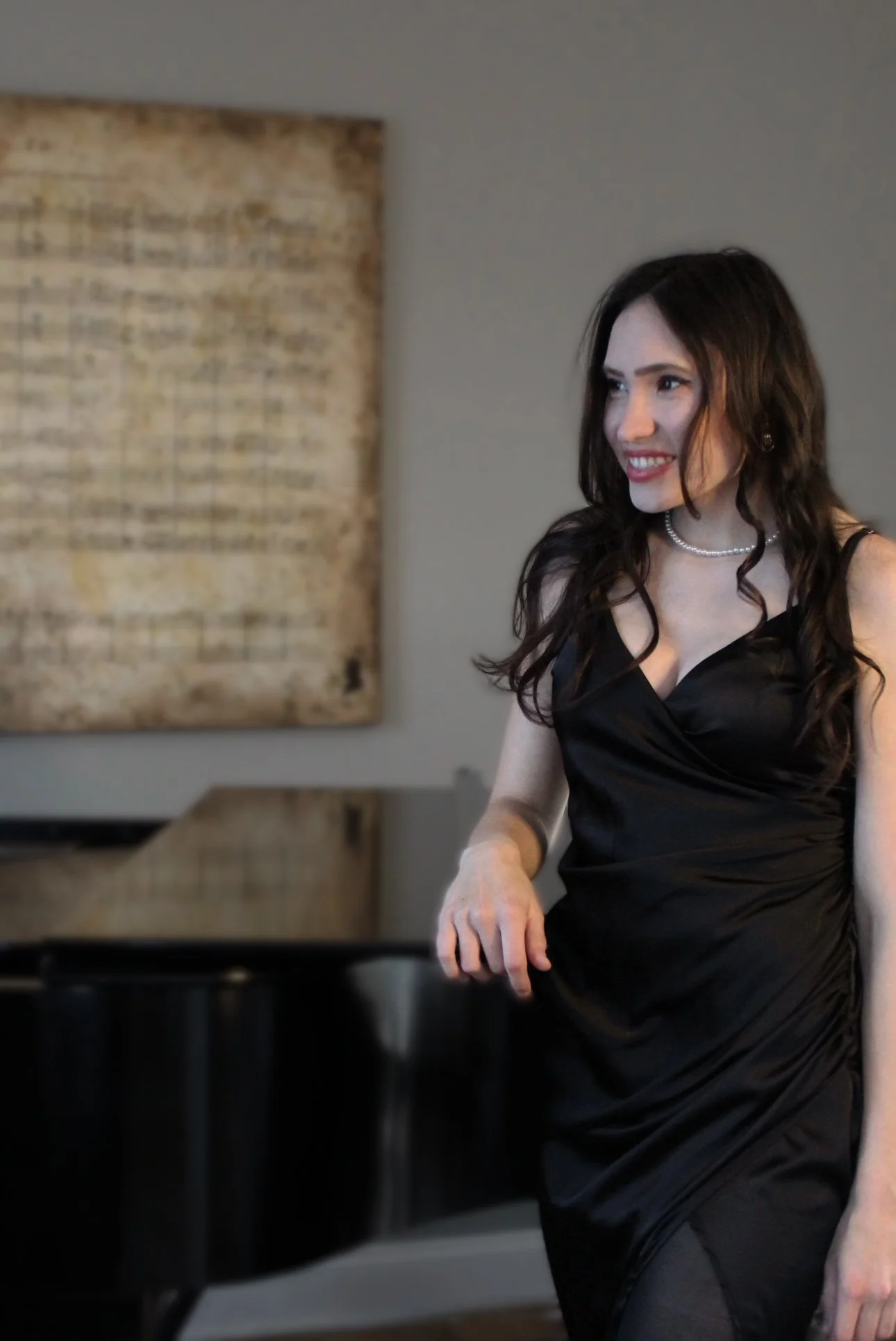 A woman with long dark hair in a black dress, smiling, standing next to a black grand piano