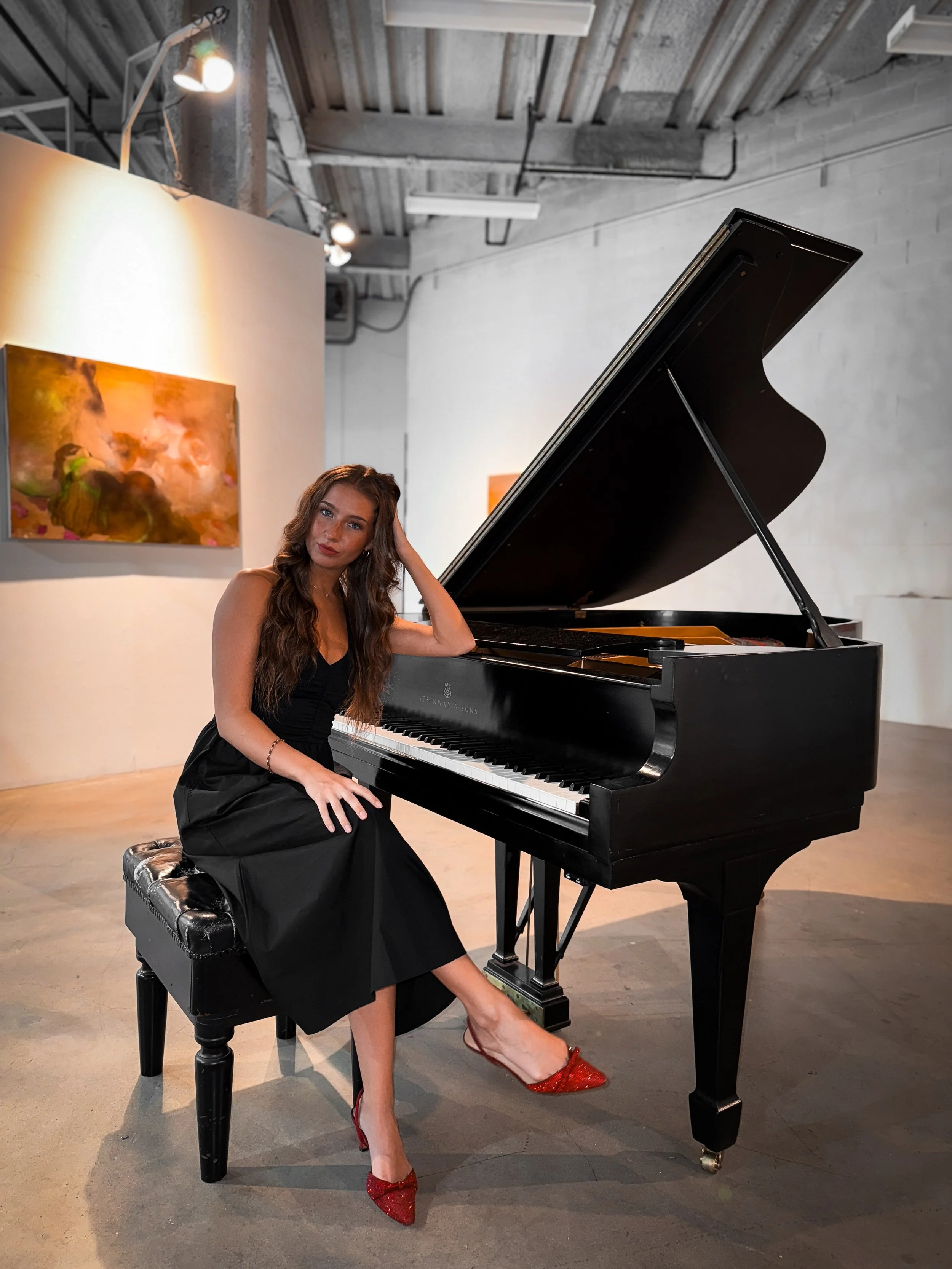 A woman in a black dress and red high heels sitting beside a grand piano in an art gallery.