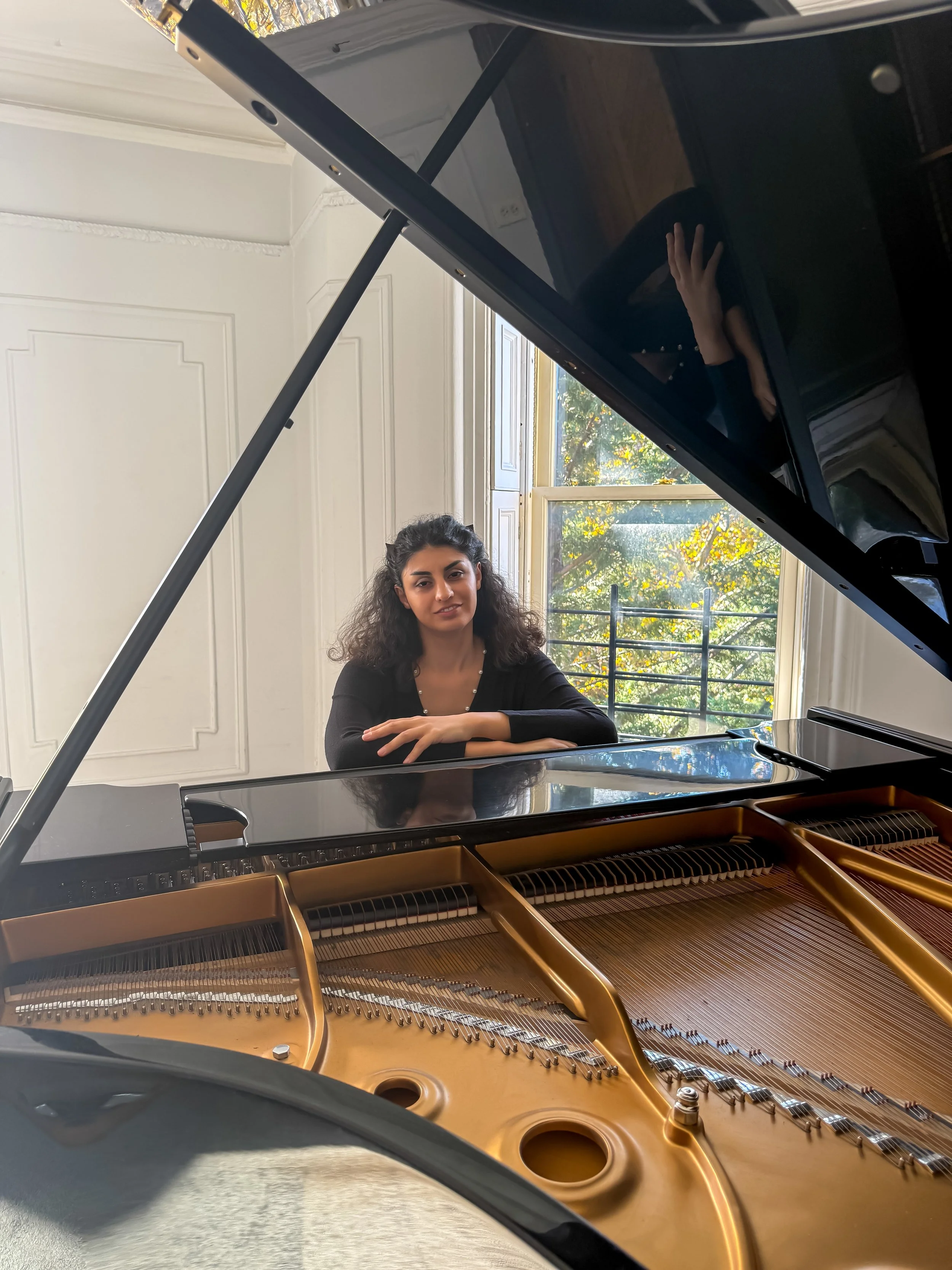 A woman with curly hair sitting at a grand piano, looking through the open lid. The room has white walls with decorative paneling and a large window showing green trees outside.
