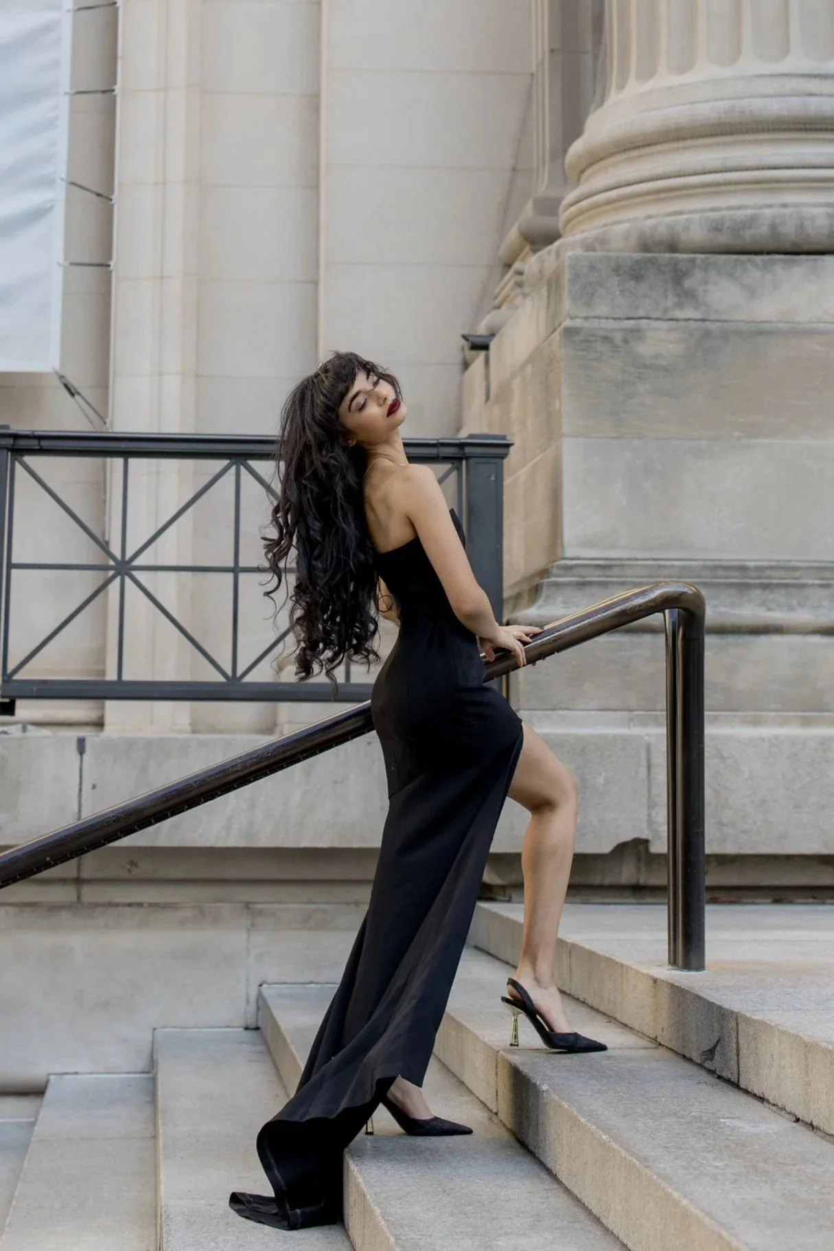 A woman with long, curly black hair and red lipstick posing on outdoor stone stairs in a black strapless dress and high heels.