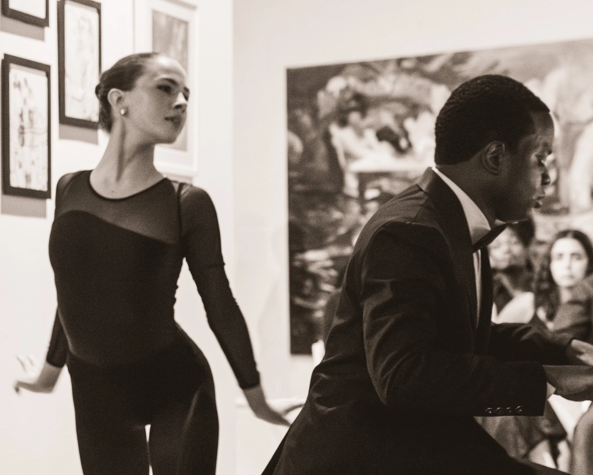 A woman in an elegant black dress with sheer details stands in an art gallery, observing artwork on the wall, while a man in a tuxedo sits nearby with his head bowed, possibly reading or writing, with other artwork visible in the background.