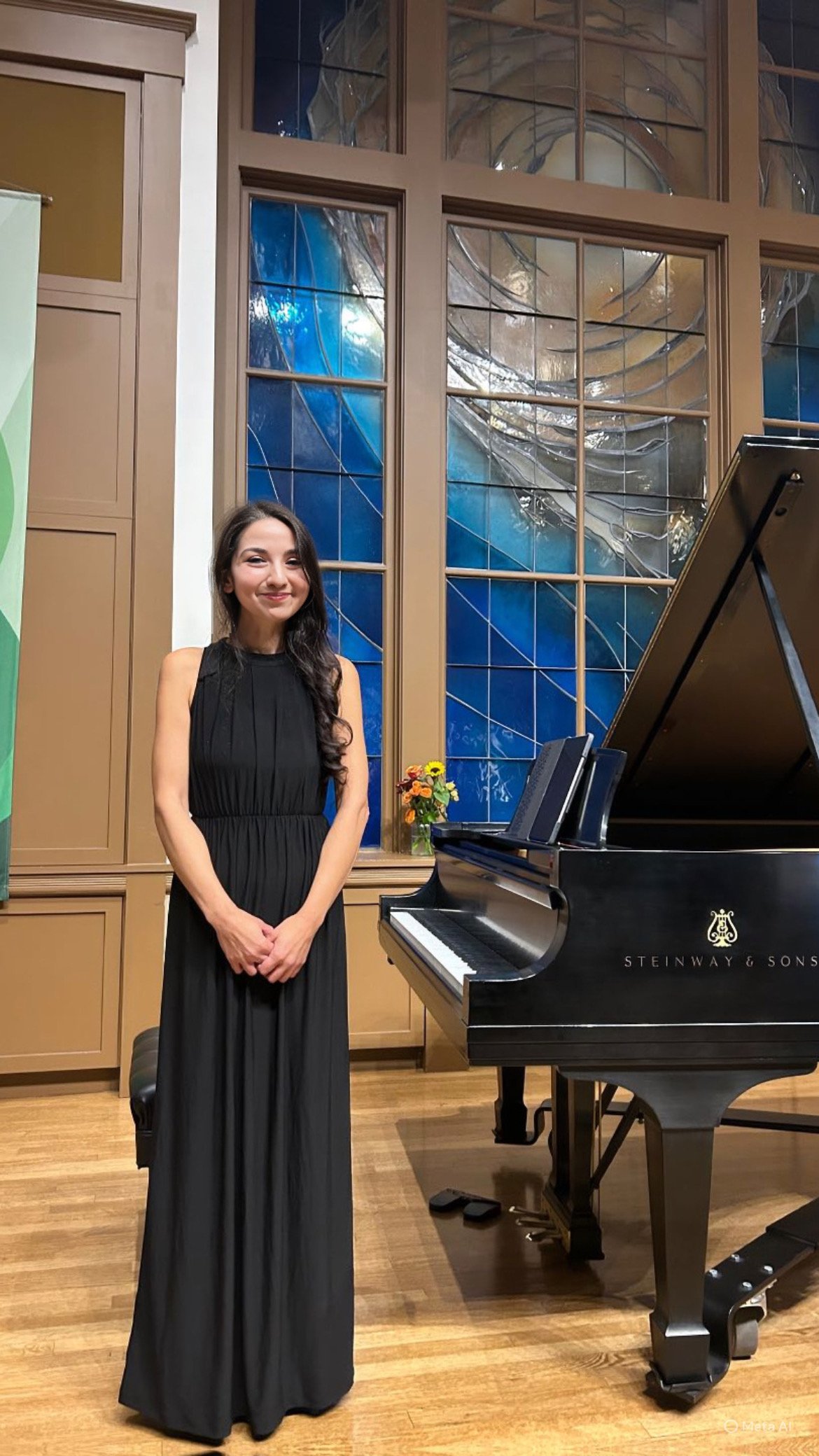 A woman in a black dress standing next to a black Steinway & Sons grand piano in a room with wooden floors and large window with stained glass design.
